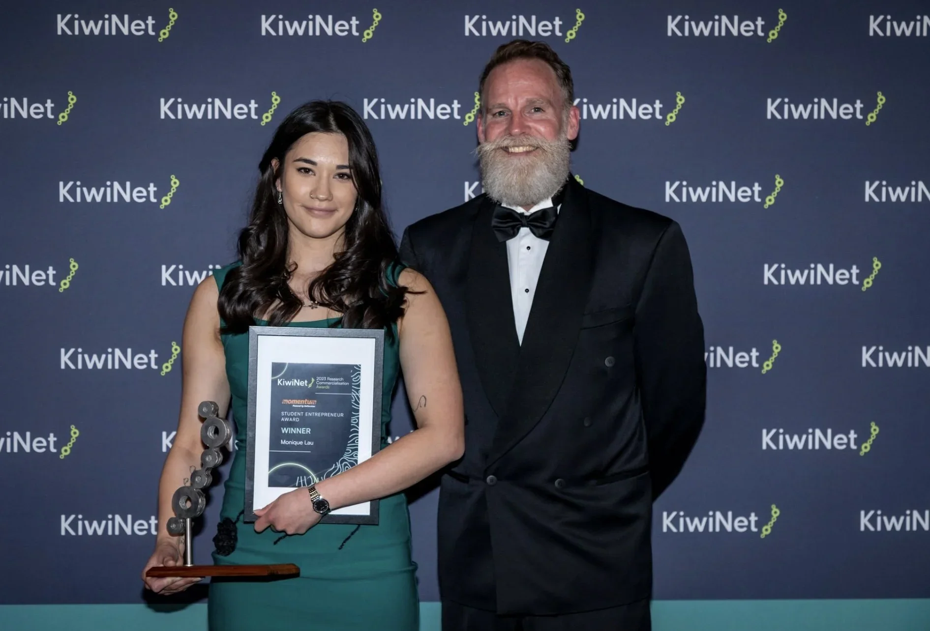A young woman in a teal dress holding an award and a trophy, standing next to a man in a black tuxedo at an event with a dark blue backdrop displaying the KiwiNet logo.