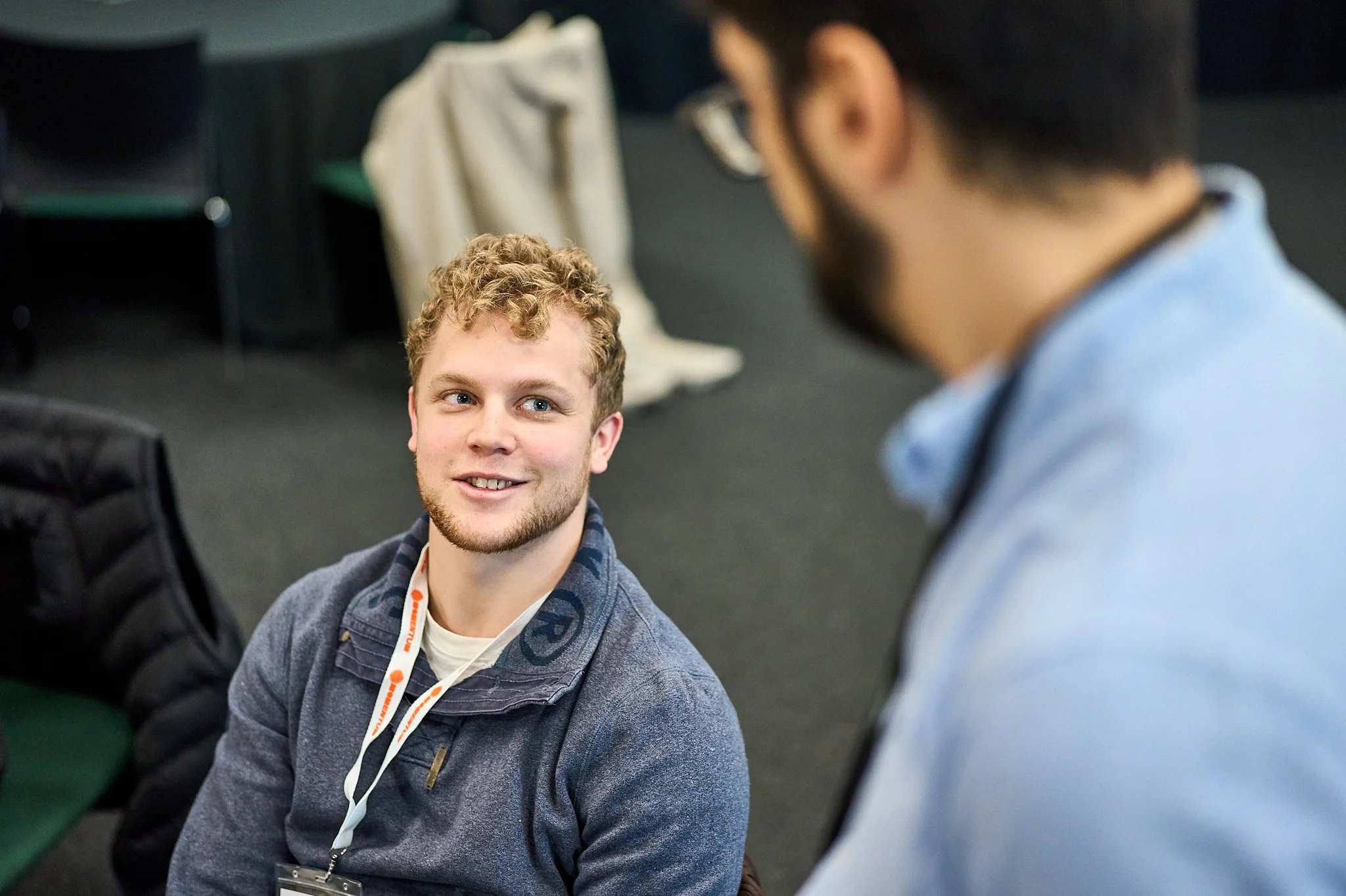 Two young men appear to be having a conversation in an indoor setting. One man is in focus, with curly blond hair, a beard, and a blue shirt, looking up and smiling. The other man, blurred, has dark hair and glasses, and is wearing a light blue jacket over a collared shirt. There are chairs and some fabric visible in the background.