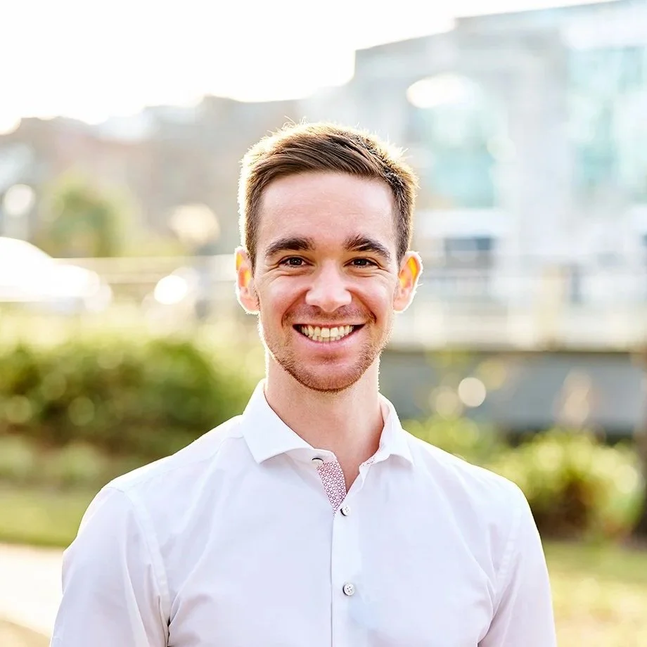 A smiling young man with short brown hair, wearing a white dress shirt, standing outdoors during daytime with a blurred background of greenery and modern buildings.