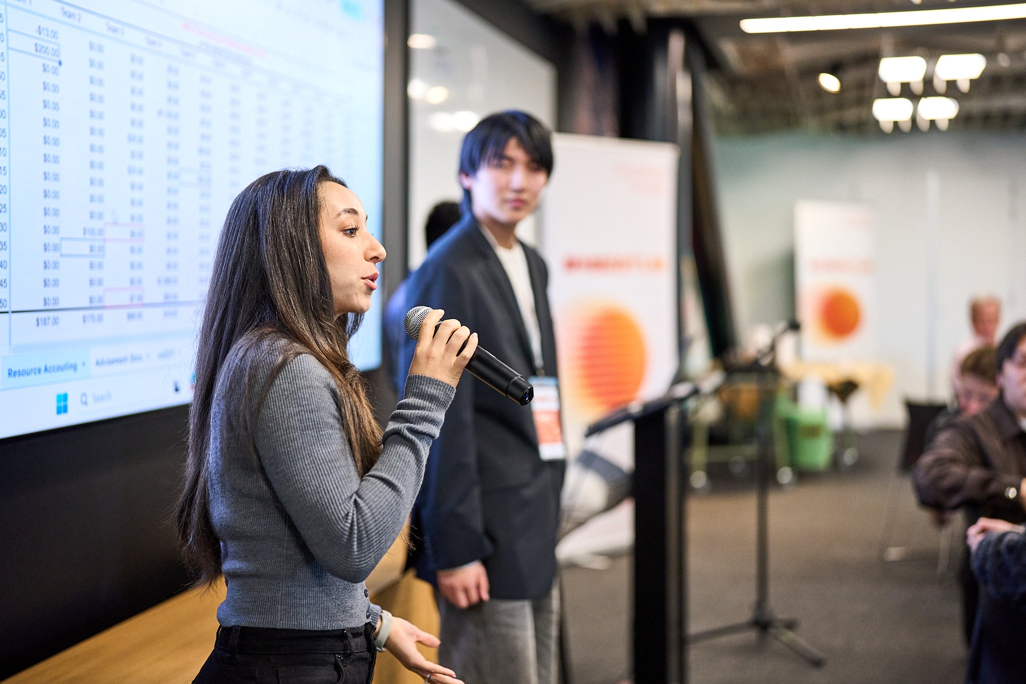 A woman speaking into a microphone in front of a large screen with data, with a man standing nearby and blurred audience in the background.