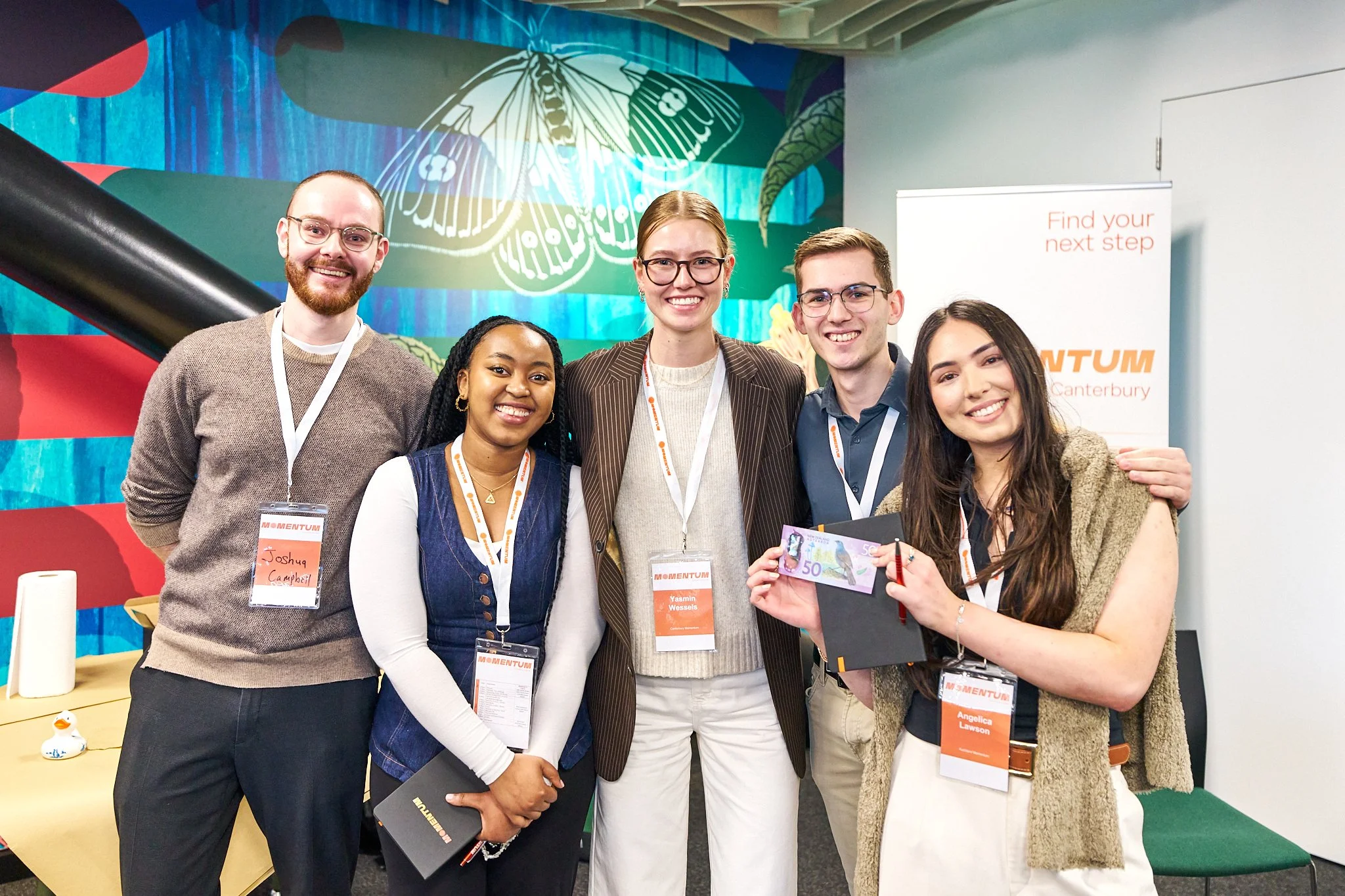 Group of five diverse young adults smiling at a conference or event, holding a fifty dollar bill, standing in front of a colorful backdrop with a butterfly mural and a banner that says 'Find your next step'.