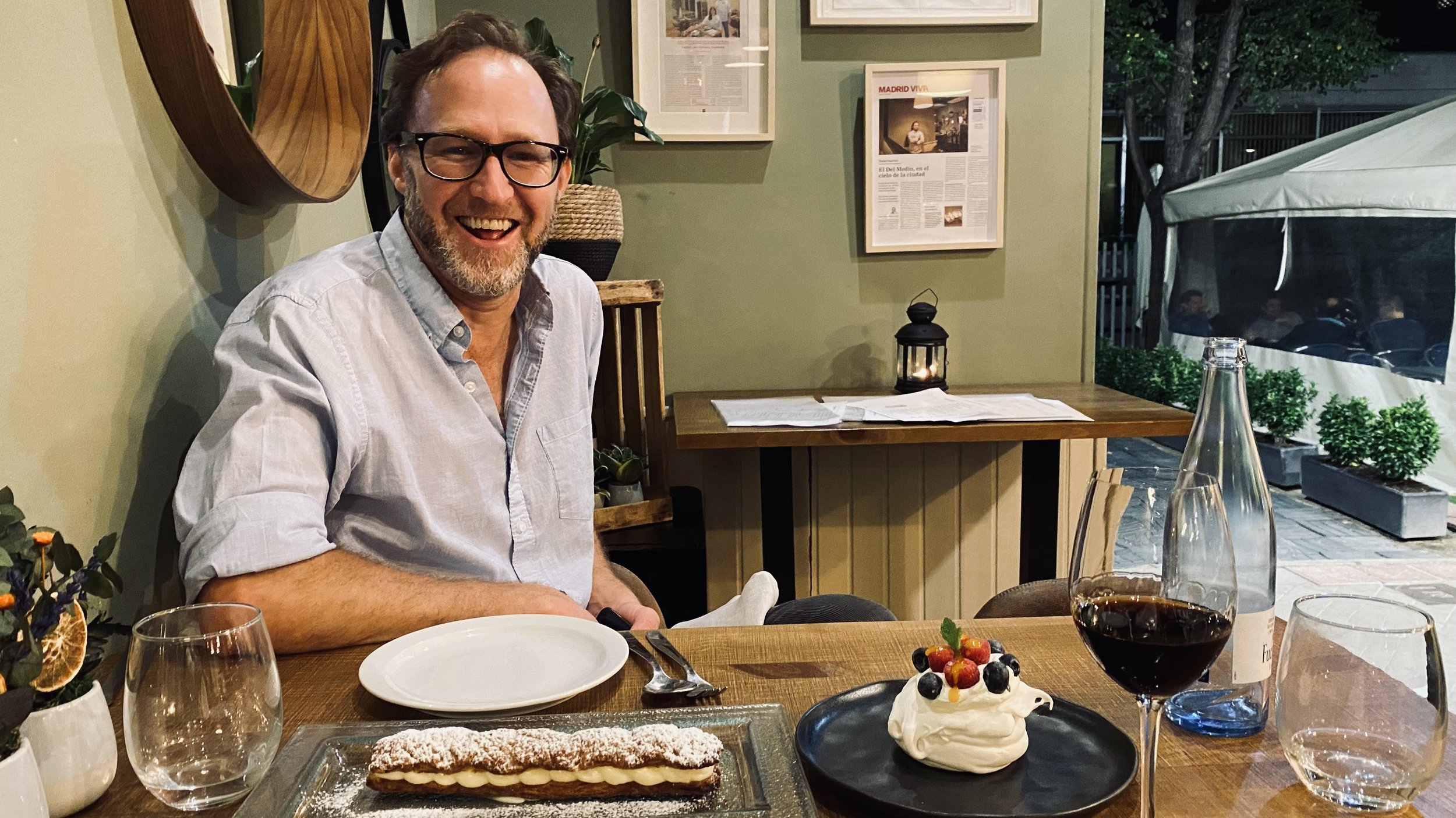A man with glasses, smiling, sitting at a dining table in a restaurant, with desserts, a glass of red wine, and a bottle of water in front of him, background includes framed pictures and a lantern candle.