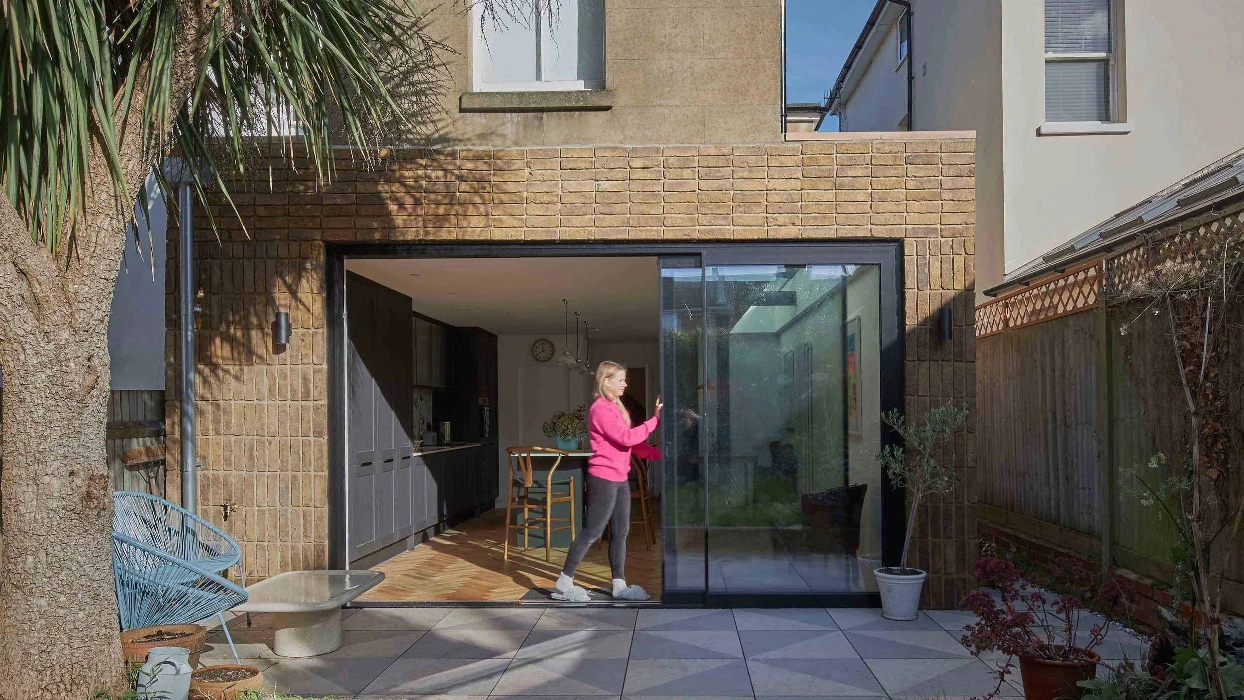 A woman in a pink sweatshirt and gray leggings standing inside a modern home, visible through large sliding glass doors, with a backyard patio and garden outside.