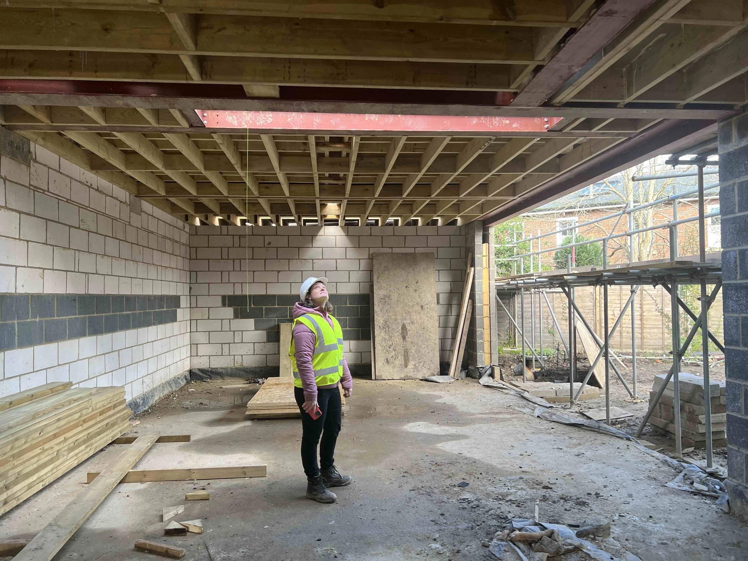 A construction worker in a safety vest and helmet inspecting the ceiling framing inside a building under construction.