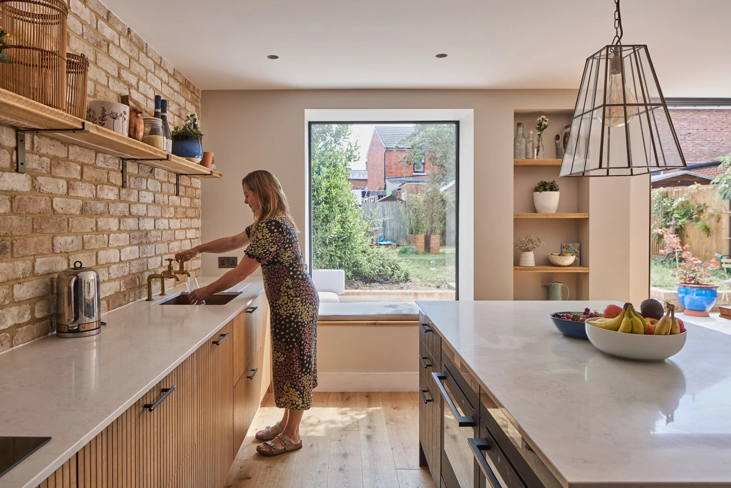 Woman washing her hands at kitchen sink with large window and fruit bowl on island in modern kitchen.