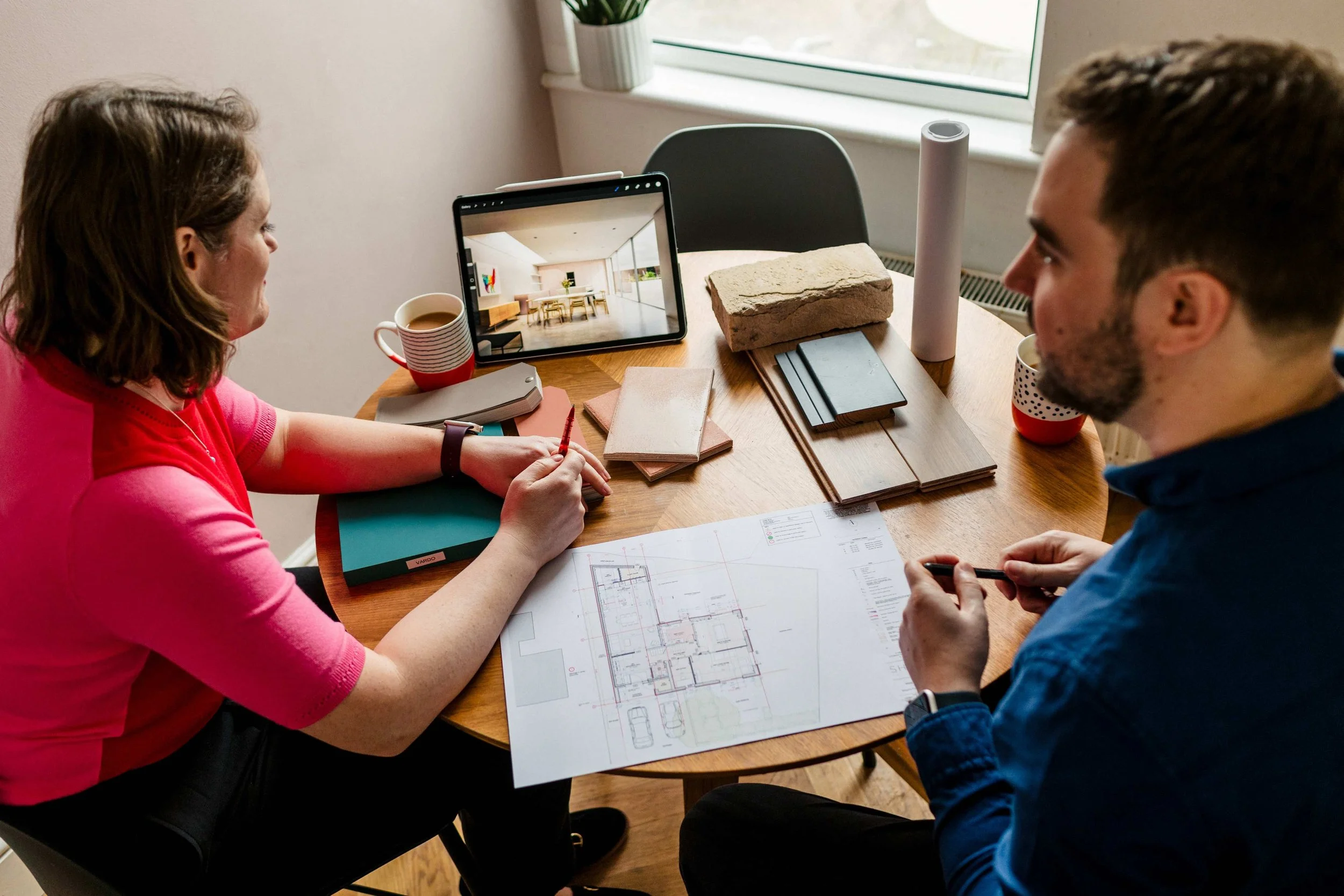 Two people sitting at a round table reviewing architectural plans, with samples and tools for interior design, a tablet displaying an empty room, and a window in the background.