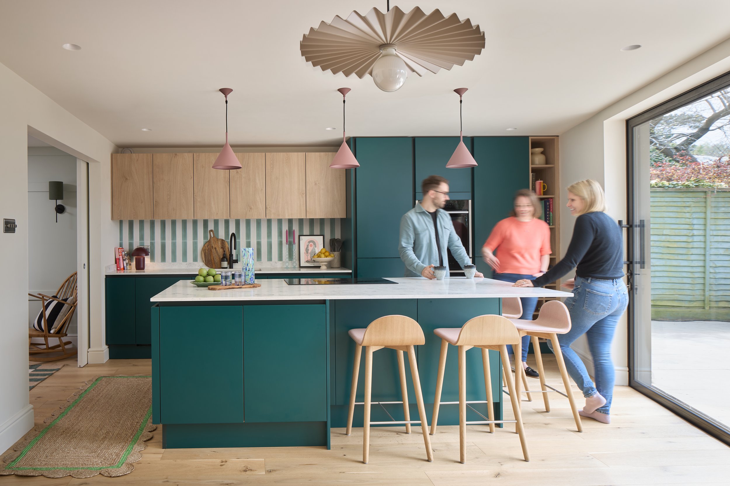 Three people with blurry faces standing and sitting at a kitchen island counter inside a modern kitchen, with large sliding glass door opening to an outdoor patio with a wooden fence and trees.