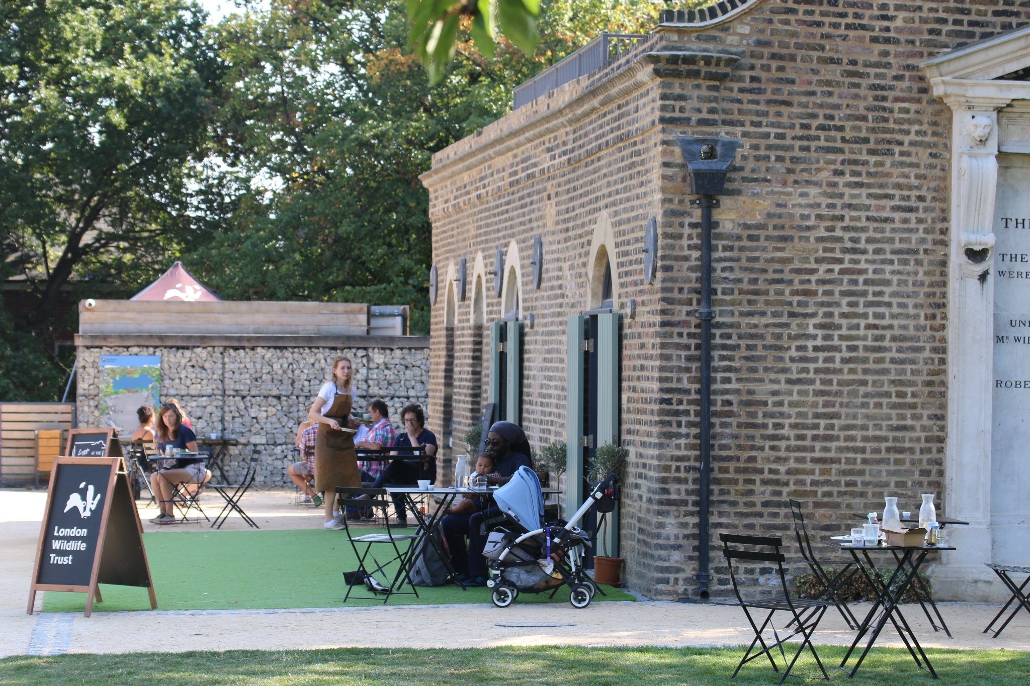 Outdoor seating area at a cafe or restaurant with tables, chairs, and a stroller. People are sitting and dining, and a waitress is taking orders. A signboard for London Wildlife Trust is visible. The background features a brick building and trees.