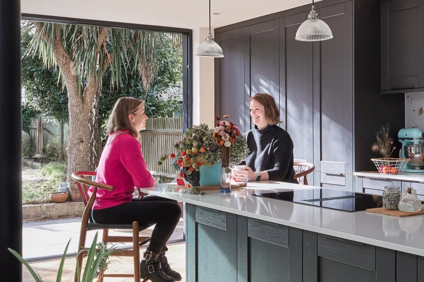Two women sitting at a kitchen island with a large flower arrangement, smiling and talking. One woman is wearing a pink sweater, the other a black sweater. There are large windows showing a backyard with trees outside.