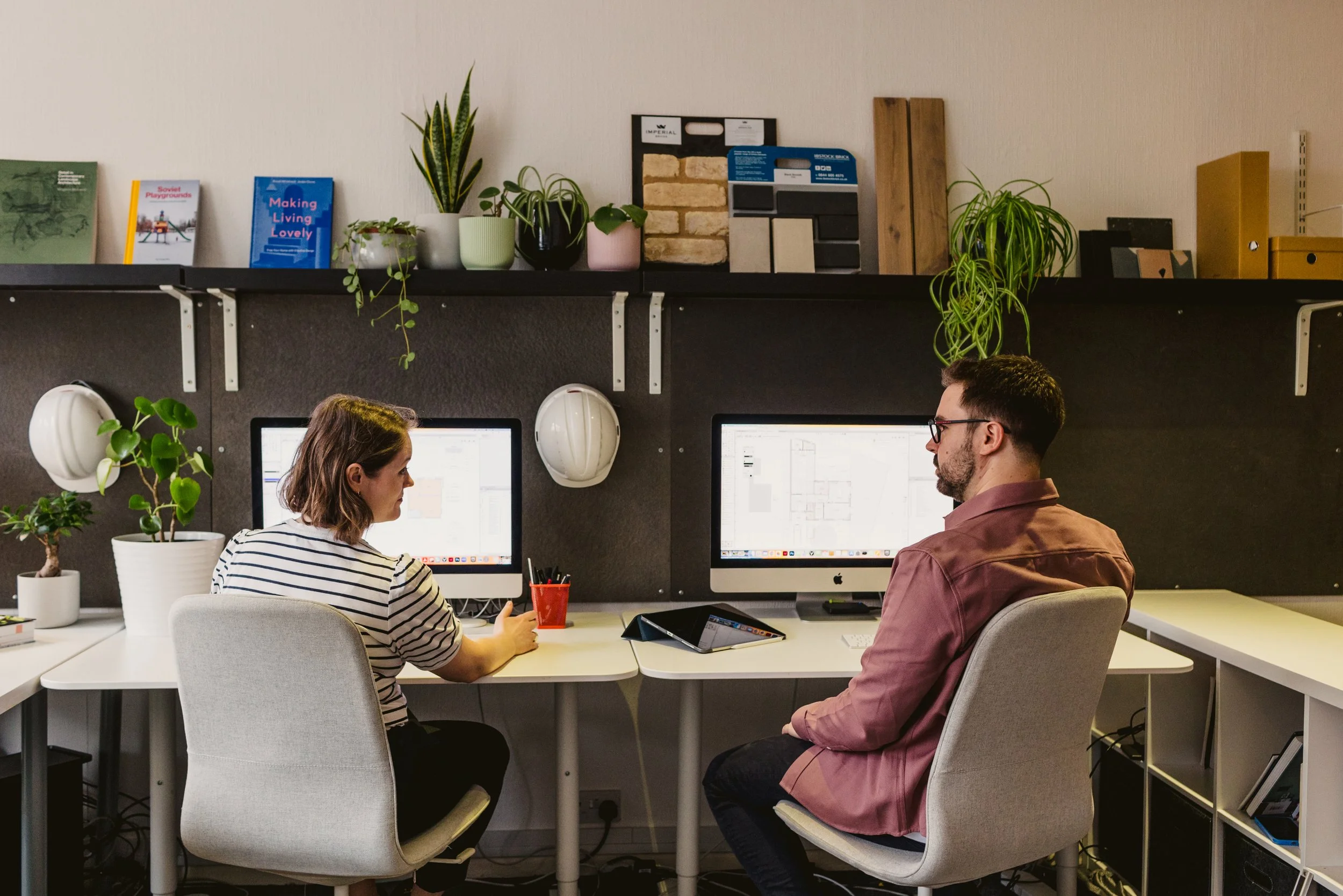 Two people sitting at a desk working on computers in an office with plants and shelves.