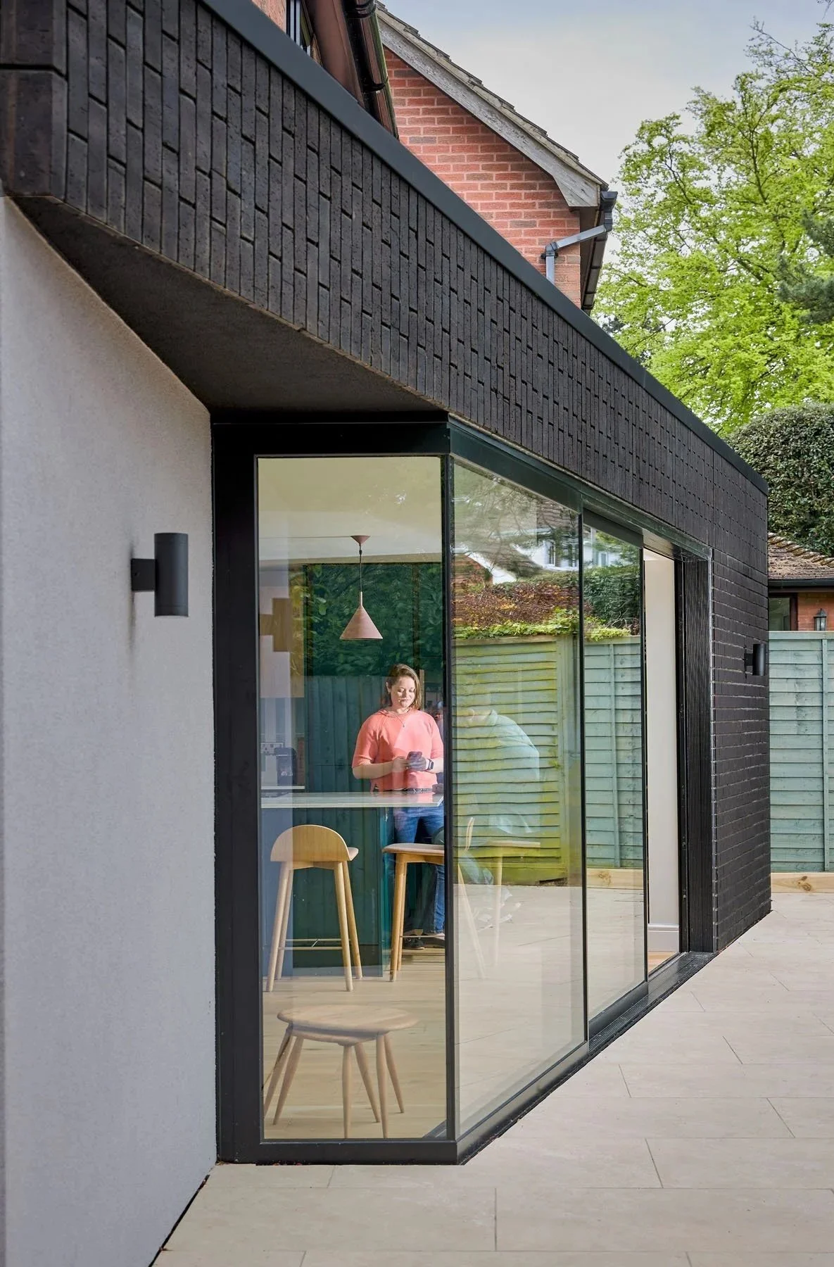 View of a modern house with large glass sliding doors, a woman inside looking at her phone, and outdoor flooring with a wooden fence and trees in the background.