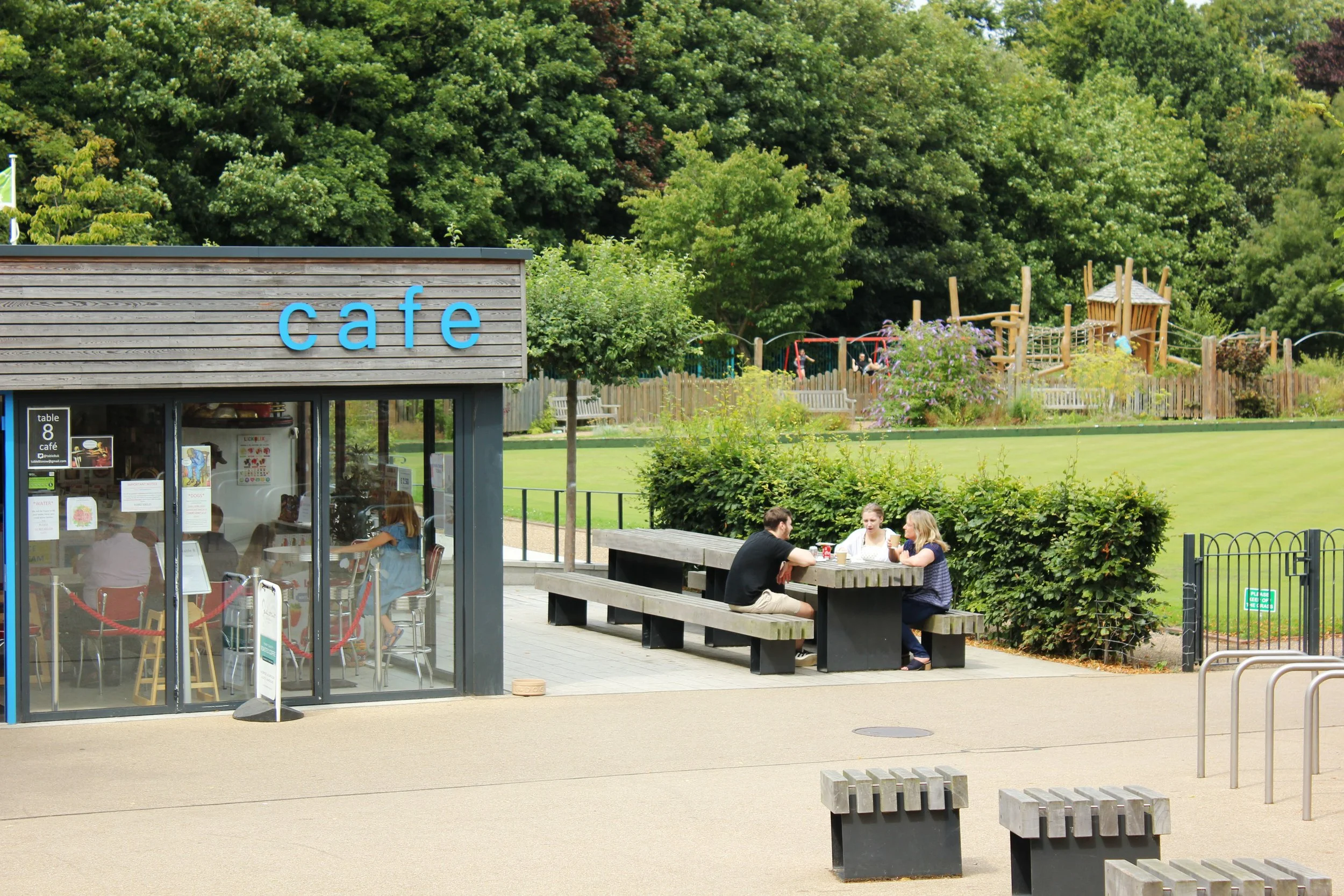 A small outdoor cafe with three people sitting at a picnic table, adjacent to a glass-fronted coffee shop, with a lush green park and playground in the background.