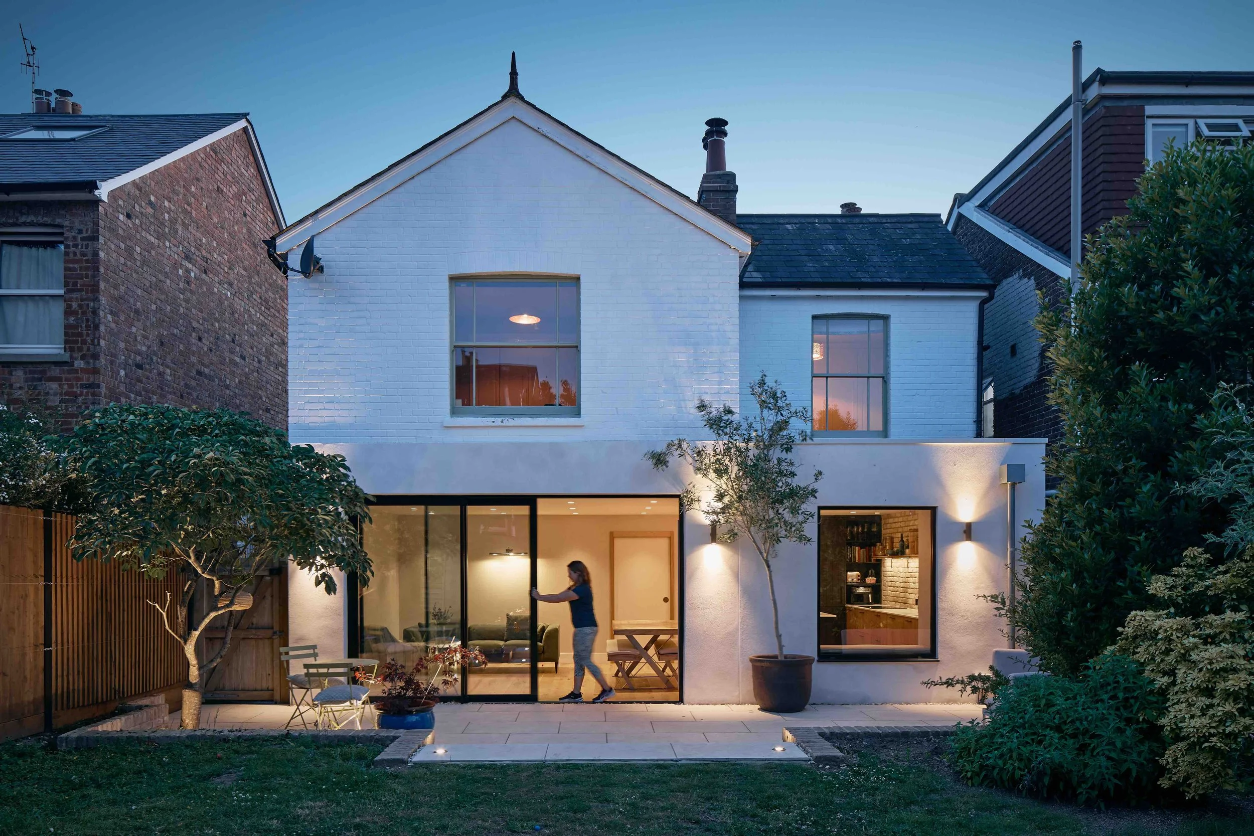 A modern two-story house at dusk with large glass sliding doors, outdoor patio furniture, and a woman opening the door. The house has a white exterior, large windows, and exterior wall lights. There are trees and shrubs in the yard.