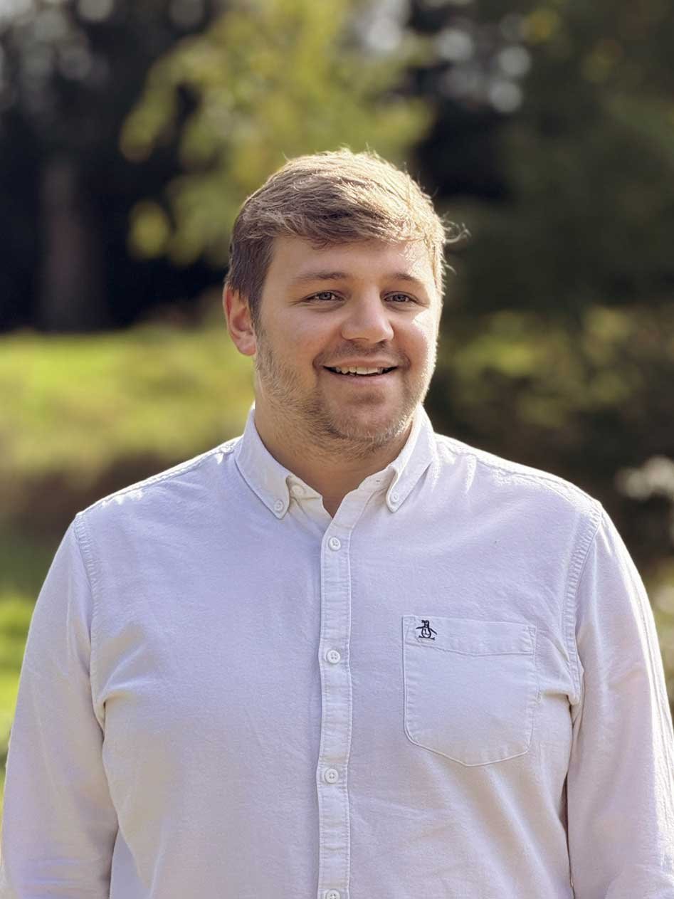 A man with light brown hair and a beard smiling outdoors, wearing a white button-up shirt with a small logo on the pocket, with a blurred background of trees and greenery.
