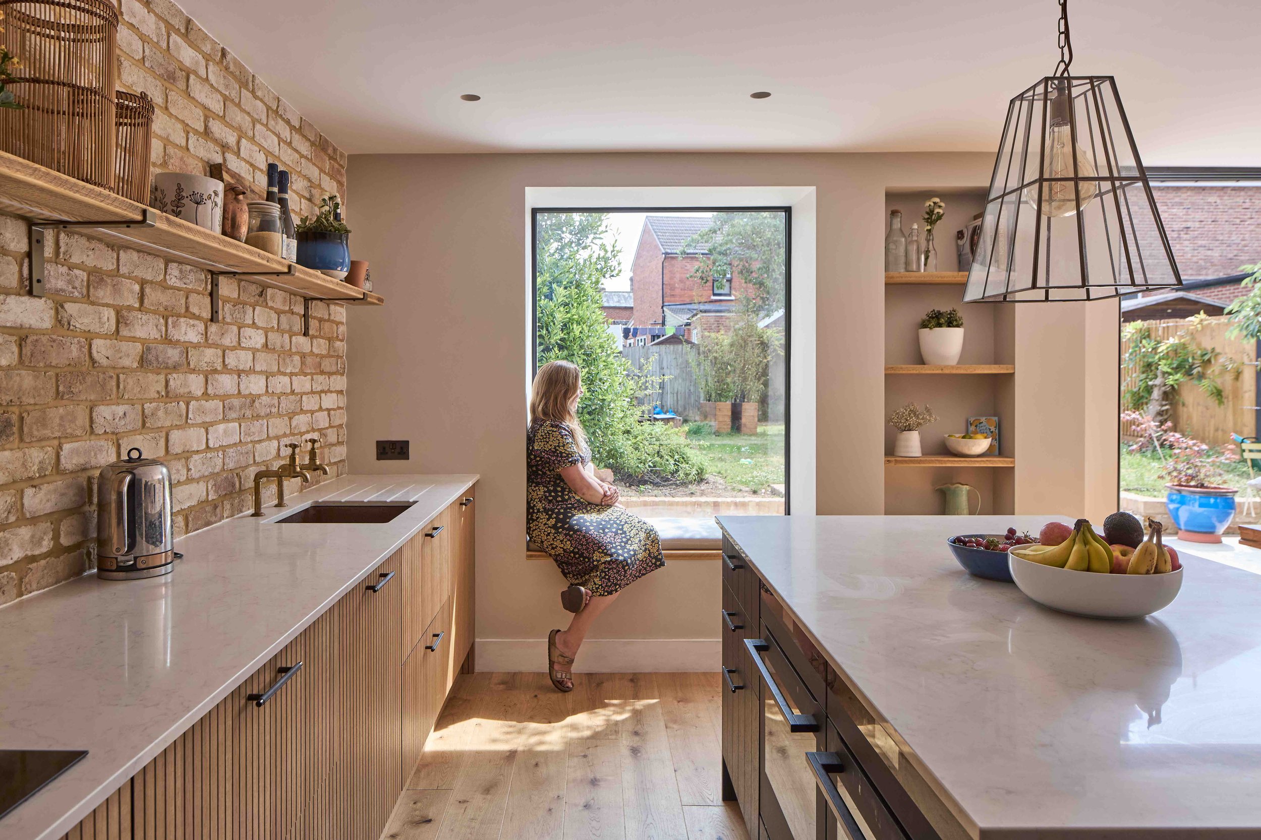 A woman sitting on a window seat in a modern kitchen, looking outside at the garden. The kitchen features a brick wall, wooden and white cabinetry, a large white marble island with bowls of fruit, and open shelves with decorative items. Natural light enters through large windows and doors leading to the garden.