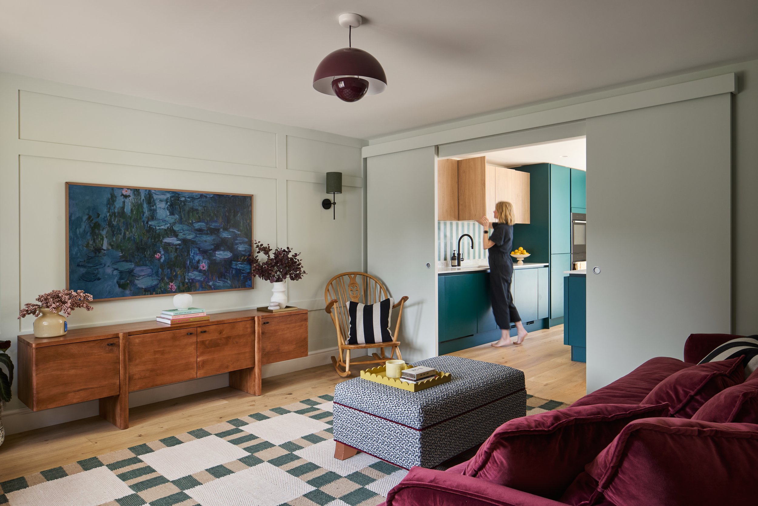 Living room with a wooden sideboard, painting of water lilies, plant pots, a chair with a striped pillow, a coffee table, and a red velvet sofa. A woman in black is in the kitchen visible through a sliding door.