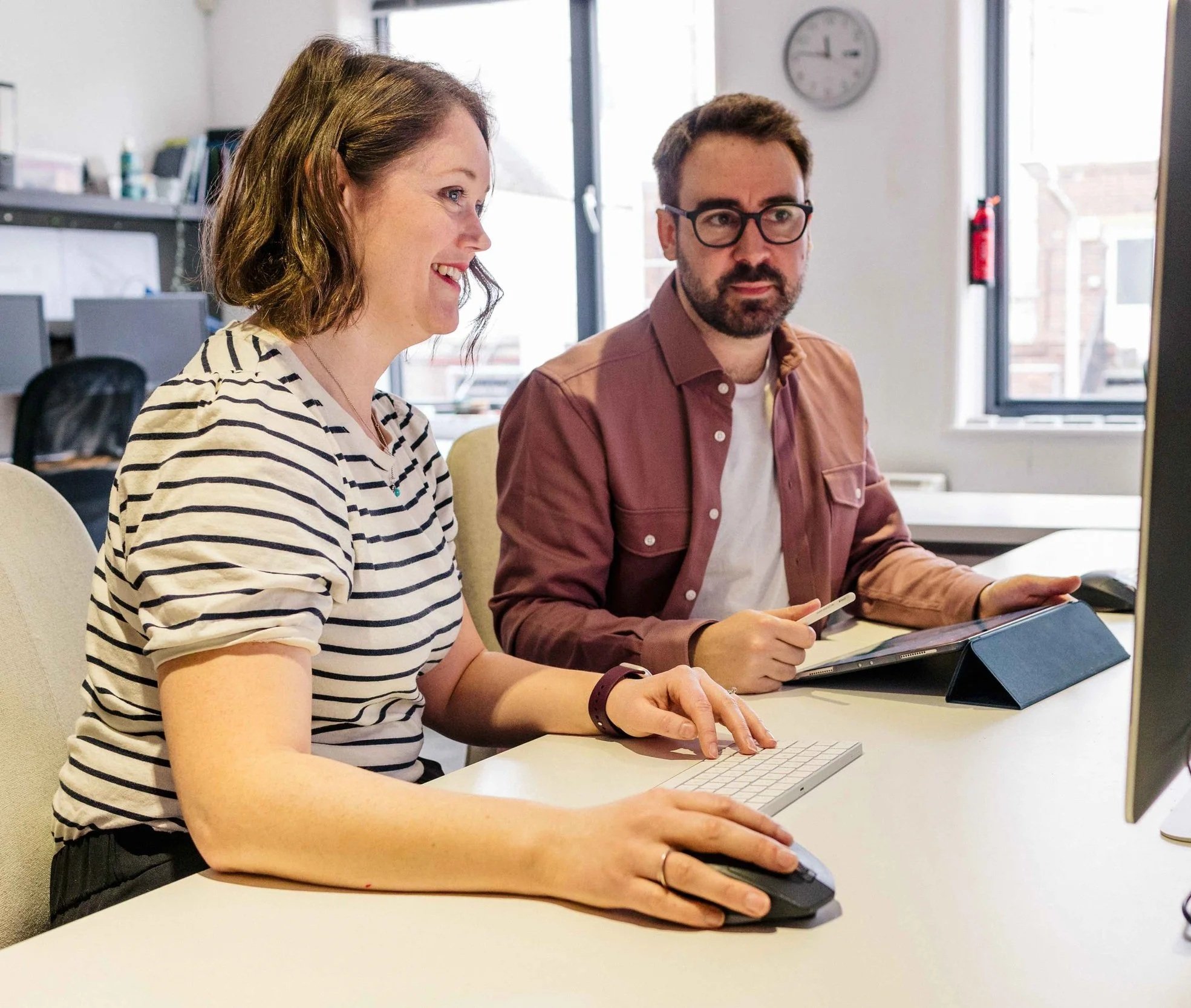A woman and a man working together at a computer in an office, with a clock on the wall and large windows in the background.
