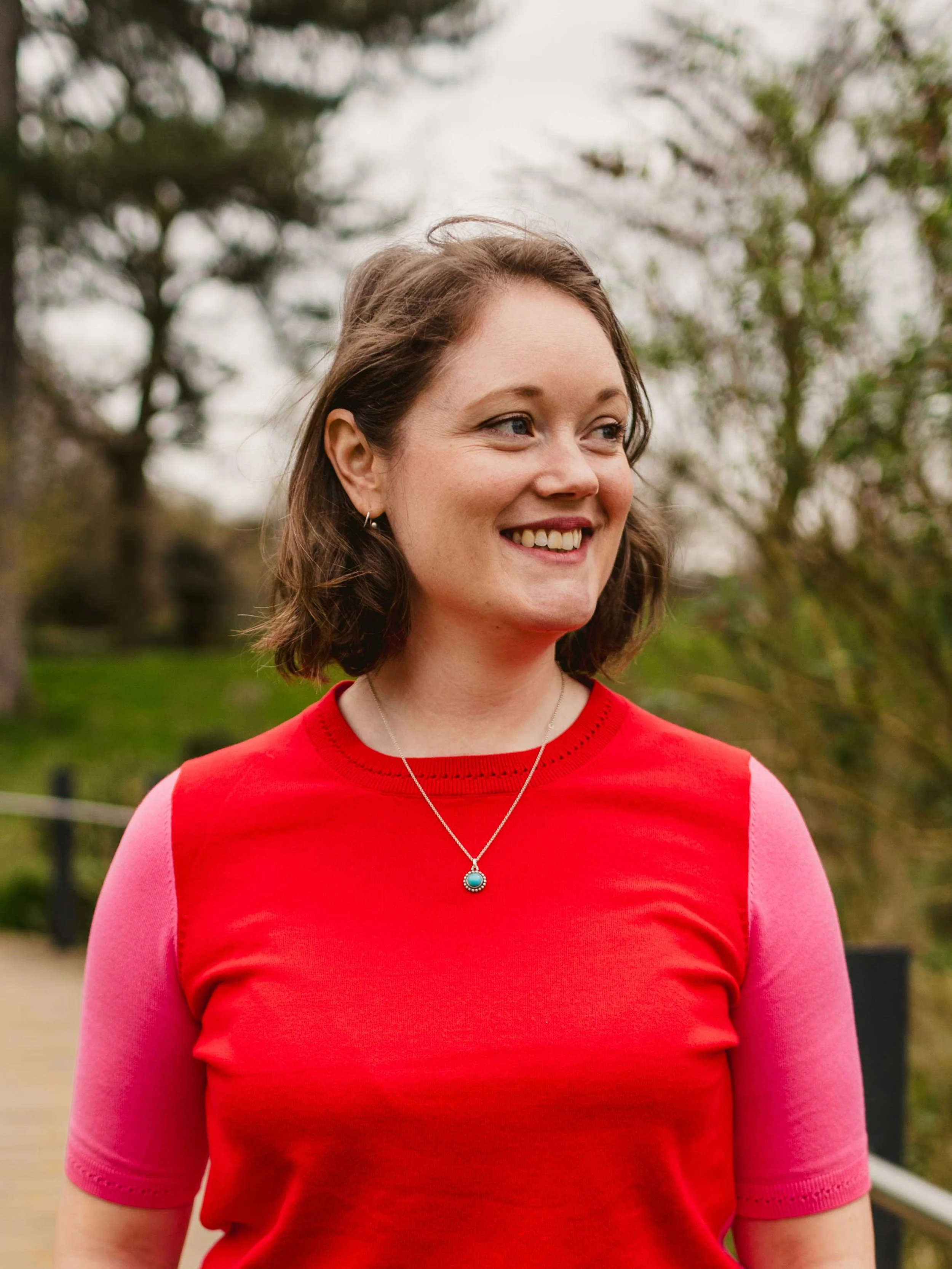 A smiling woman with shoulder-length brown hair, wearing a red and pink shirt and a silver necklace outdoors, with trees and greenery in the background.