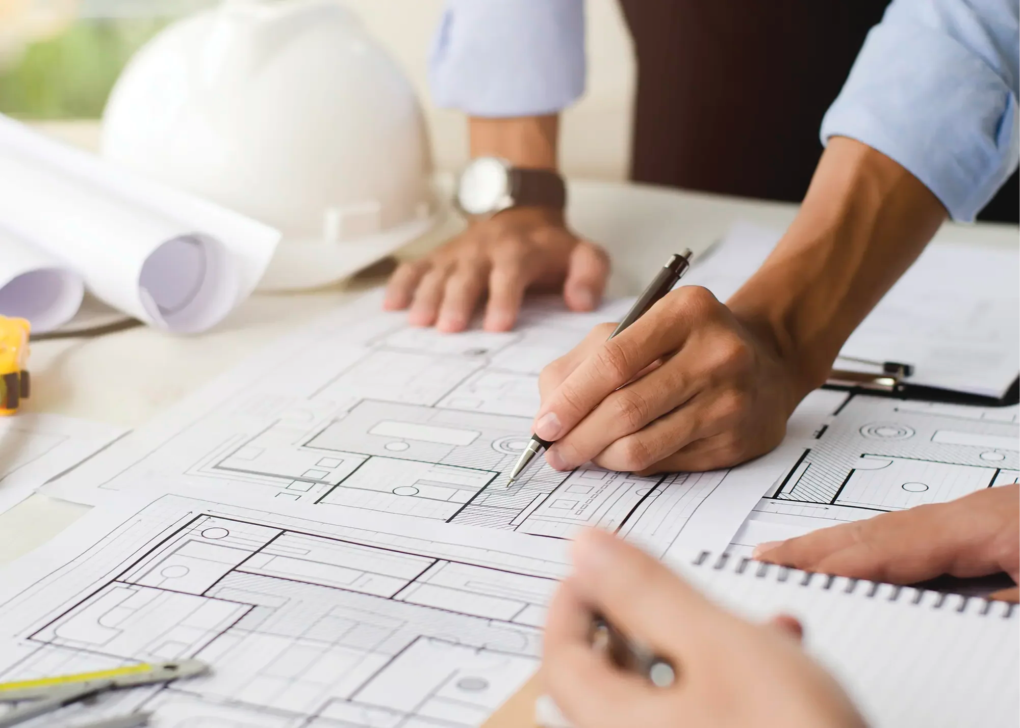 Architects working on blueprints at a construction table, with a safety helmet and rolled-up plans nearby.