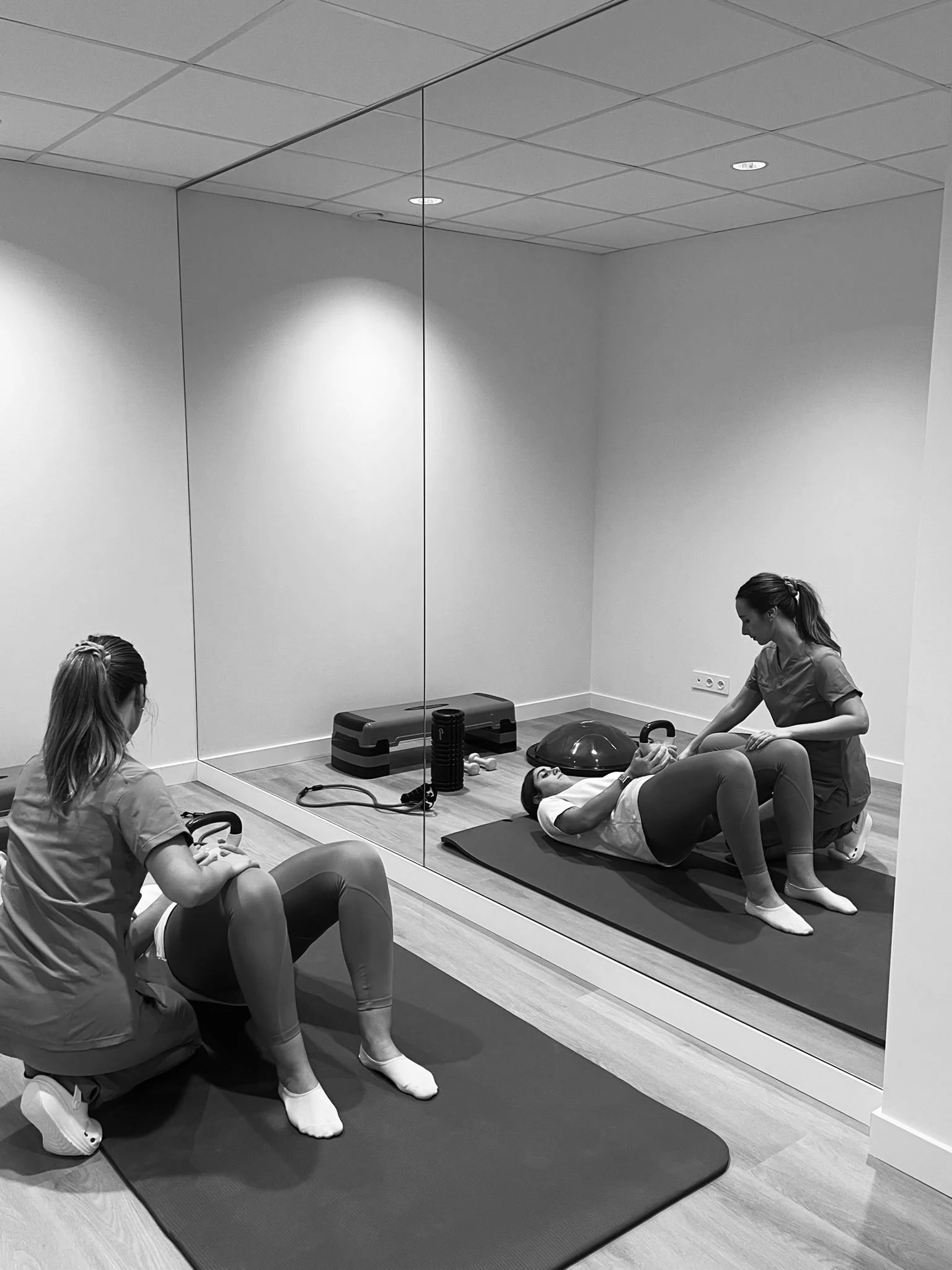 Dos fisioterapeutas asisten a una mujer que realiza ejercicios en un gimnasio de rehabilitación, con equipos de apoyo y colchonetas.