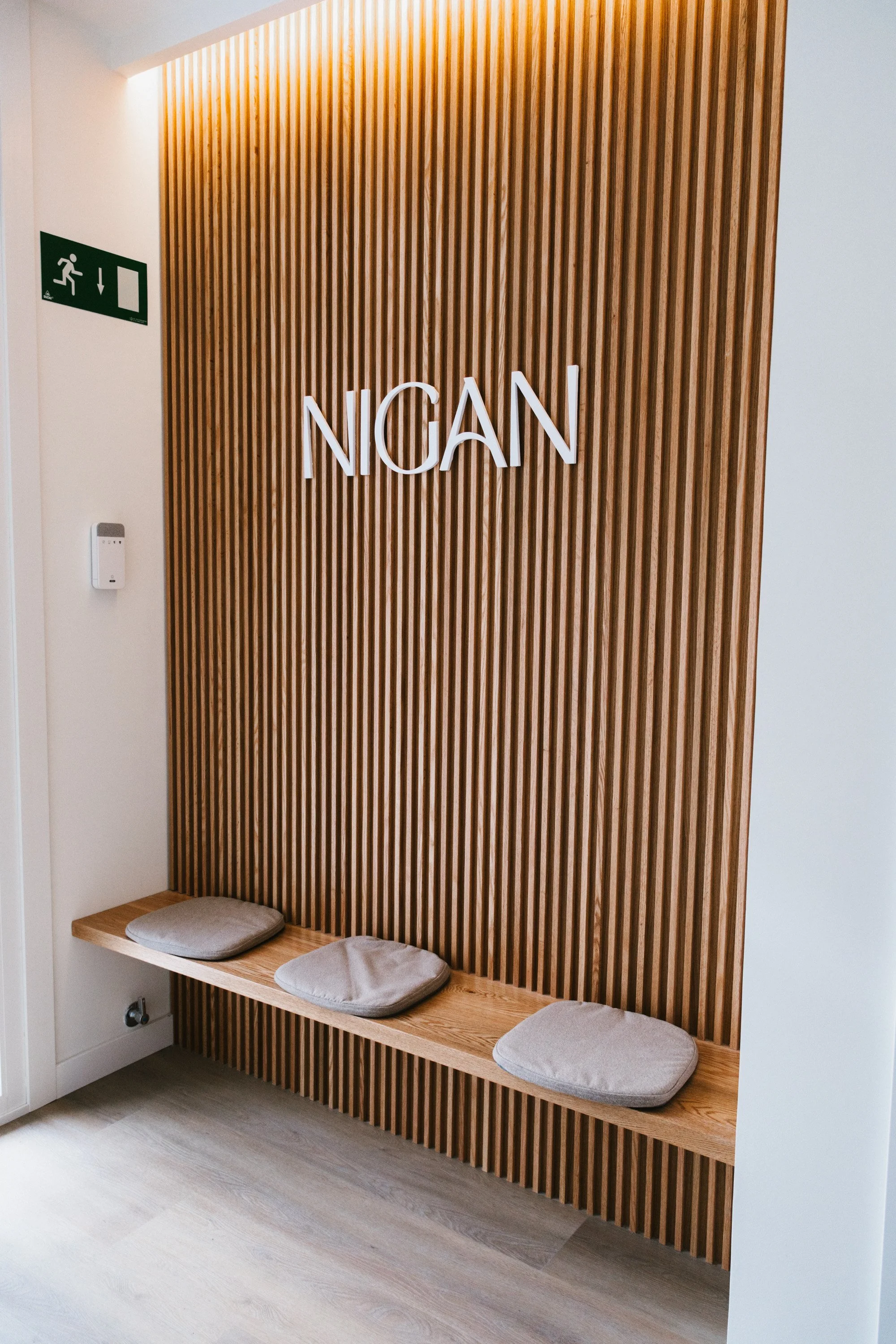 Mural de madera con la palabra 'NGAN' en letras blancas. Tres cojines gris en un banco de madera debajo del mural, esquina de una sala moderna, señal de salida de emergencia visible en la pared izquierda.