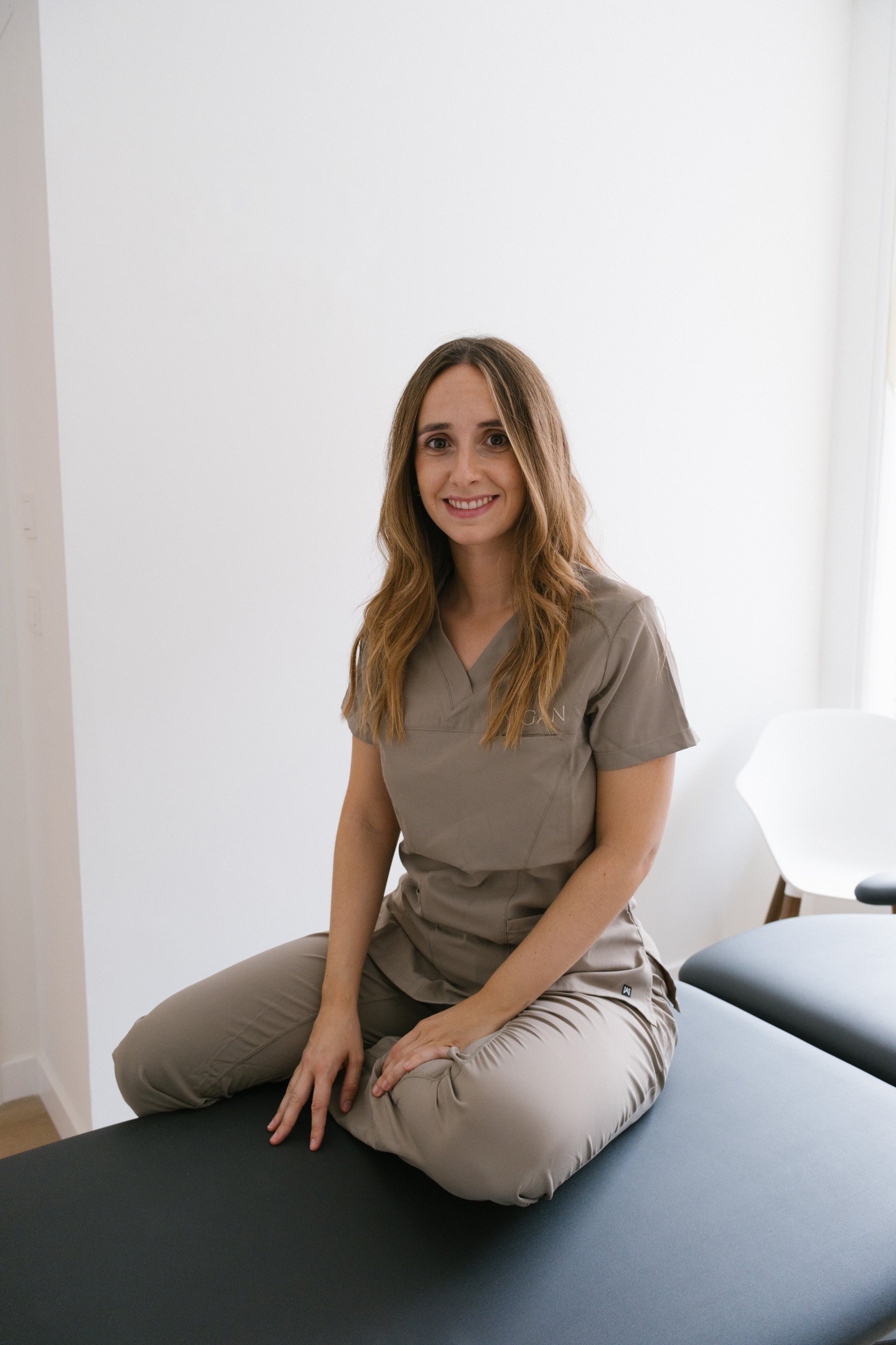 Mujer con uniforme de trabajo en una clínica, sonriendo y sentada en una camilla o mesa de exploración.