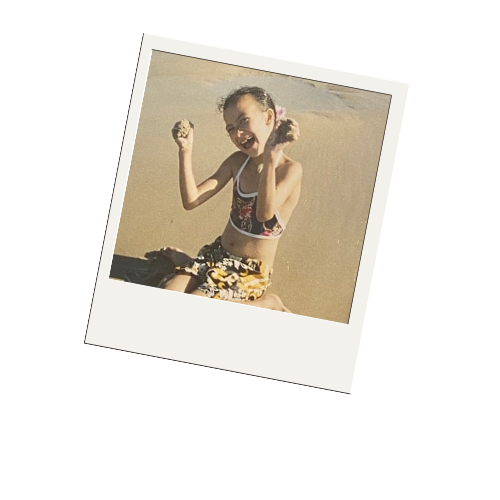 A young girl at the beach wearing a black swimsuit with floral patterns and yellow shorts, smiling and making fists with her arms raised.