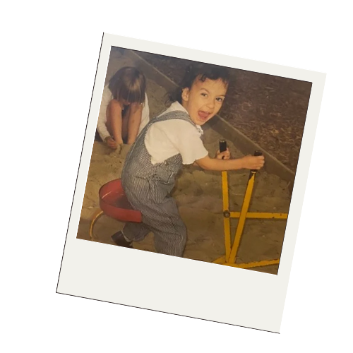 A young boy playing outdoors, sitting on a red stool, smiling and holding onto a yellow balancing beam. A girl is in the background, engaged with something on the ground.
