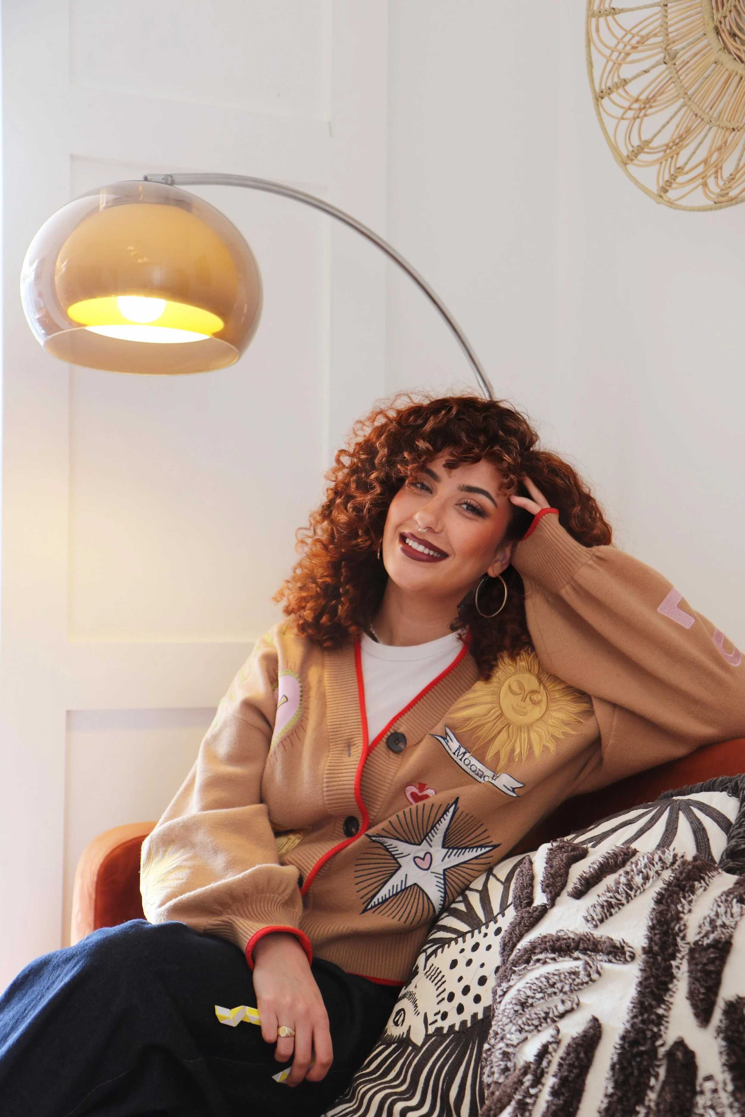 A woman with curly red hair and hoop earrings, smiling and sitting on a couch in a room with white walls, a gold desk lamp hanging overhead, and decorative pillows.