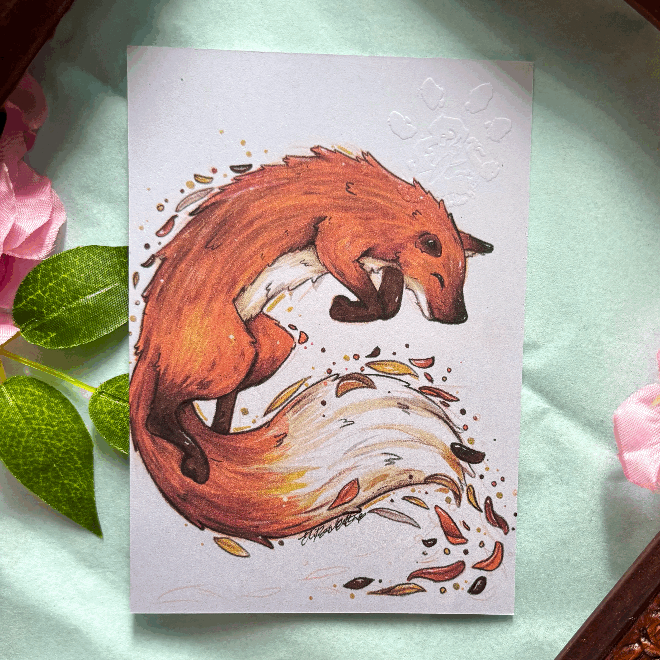 A colorful illustration of a fox curled into a spiral, with its tail forming a whirl. The fox appears to be looking down at a swirling pattern that includes leaves and small seeds around it. The artwork is detailed with shades of orange, white, and black, and is set against a white background with decorative embossed elements.