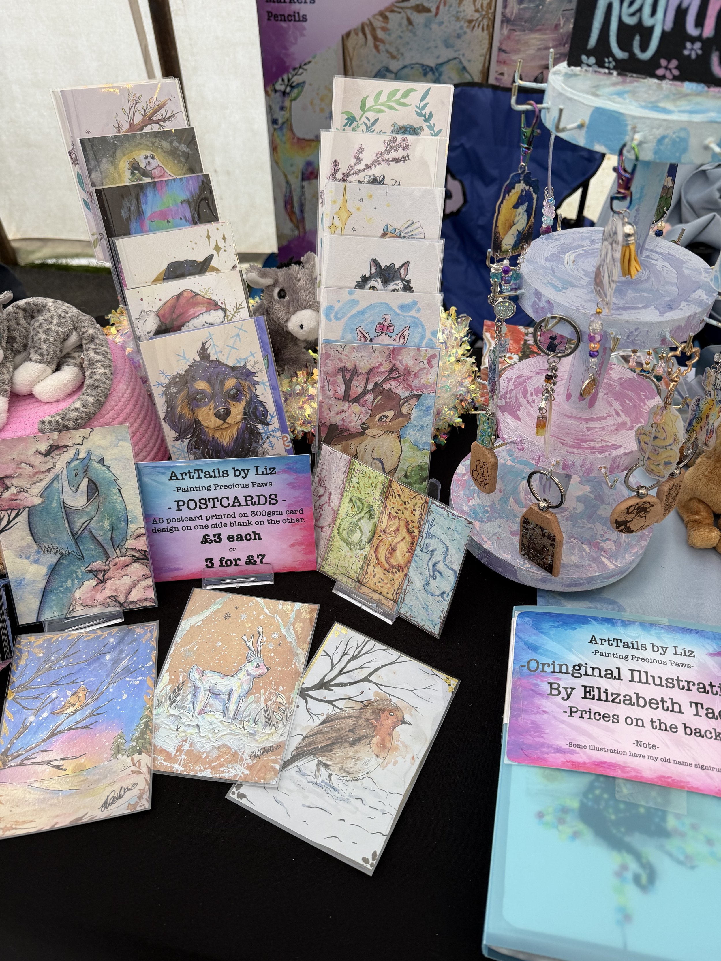 Display of illustrated postcards and jewelry at an art market booth, featuring animal and nature designs, including a deer, wolf, and birds, with a sign indicating prices and artist information.