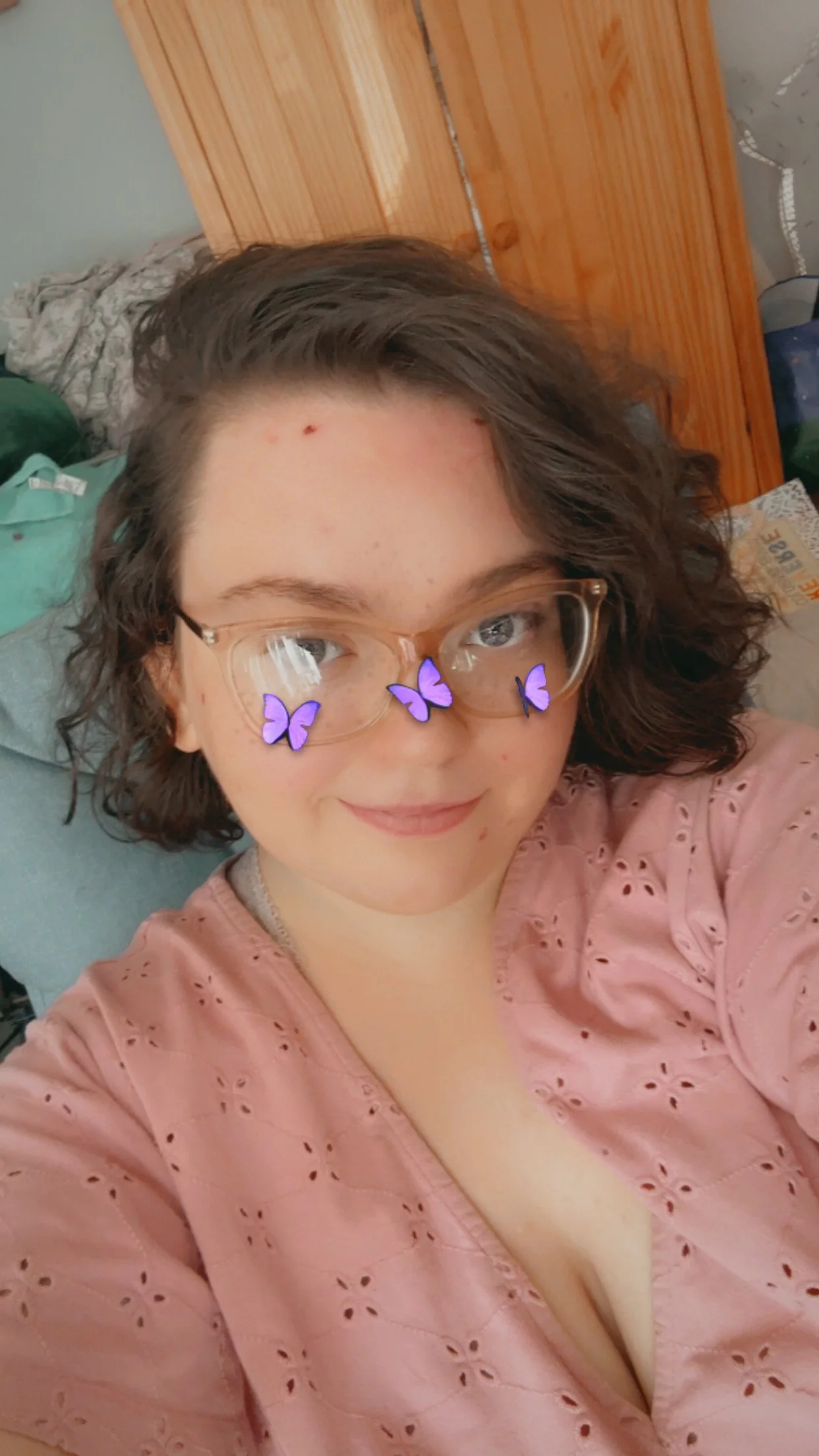 A woman with short curly hair and glasses taking a selfie in a room with a wooden dresser behind her. She is wearing a pink top and has purple butterfly filters on her glasses.