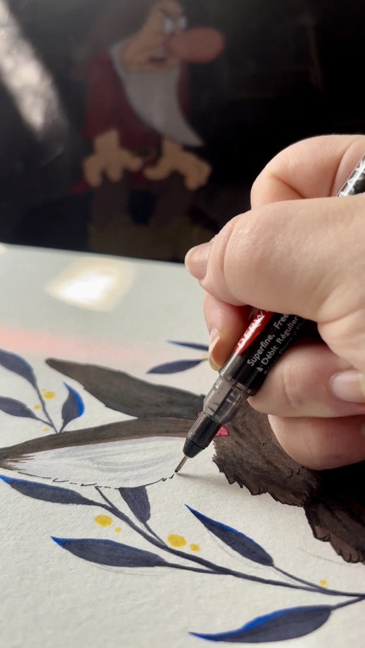 Close-up of an artist's hand drawing with a fine black pen on a sheet of paper, featuring a botanical illustration with leaves and small yellow dots, with a blurred illustration of a woman in the background.