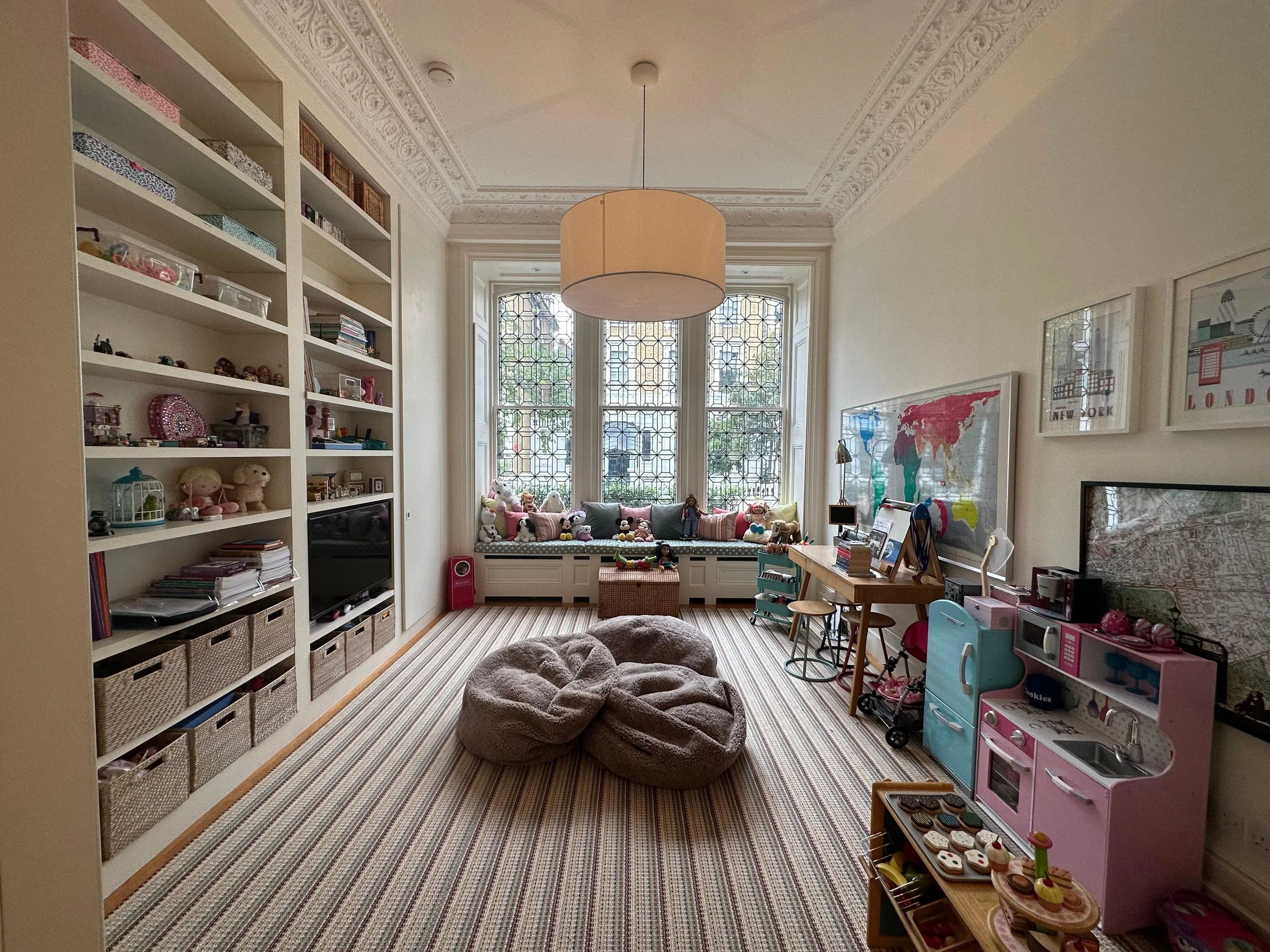 Bright children's playroom with large bay window, bookshelf filled with toys and books, plush bean bag chairs, and play kitchen set.