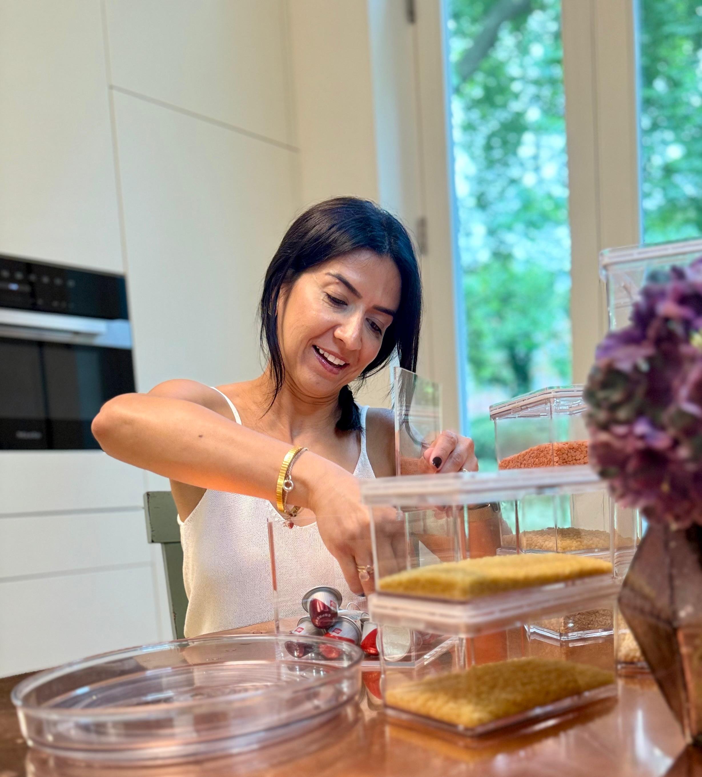 A woman with dark hair, wearing a white sleeveless top, preparing or organizing items in a clear plastic container in a bright kitchen with a window and green trees outside.