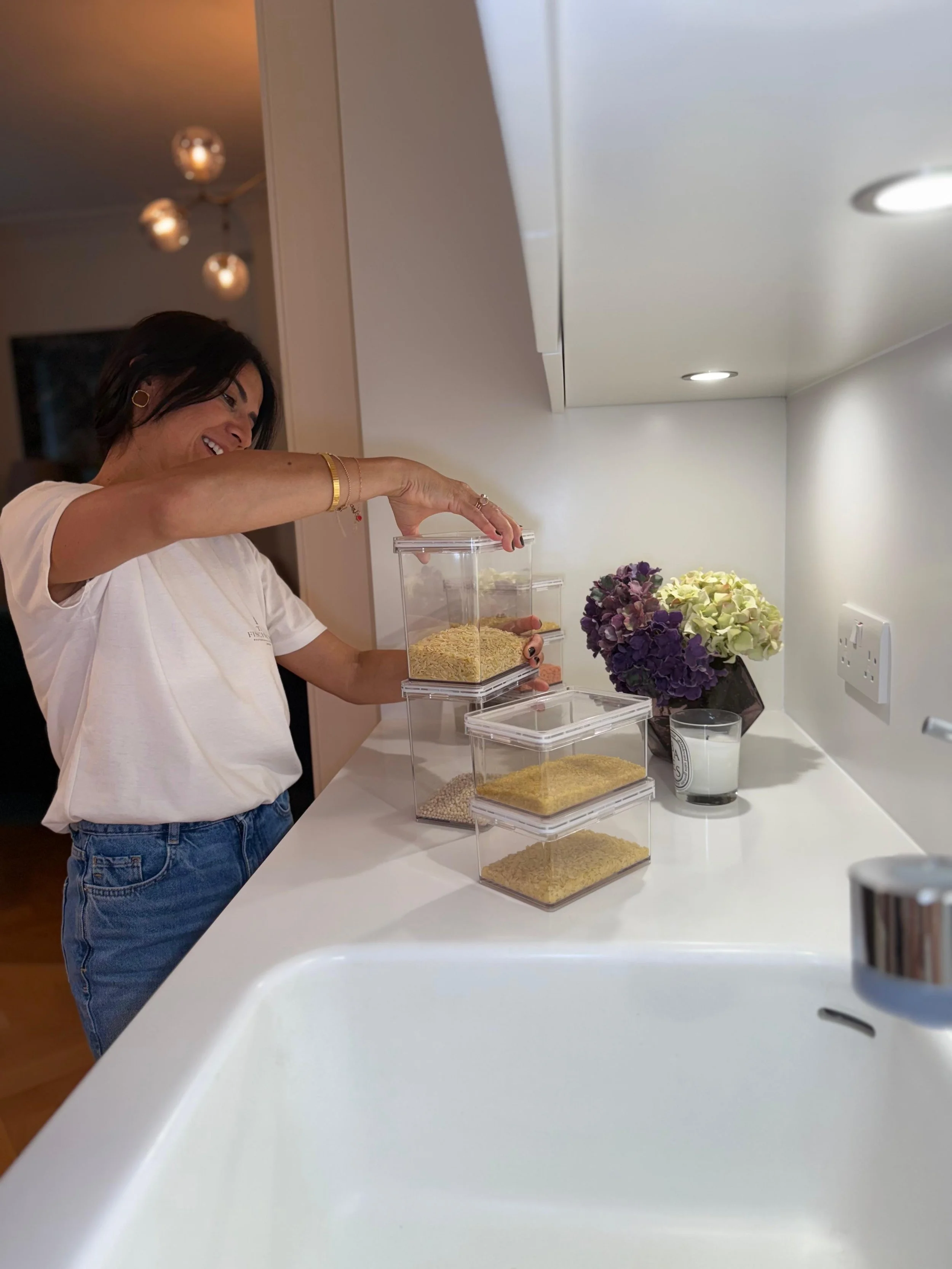 A woman smiling and scooping dry food or grains from a plastic container into another container on a white kitchen countertop. There is a vase of purple and white flowers, a lit candle, and a white wall socket nearby.