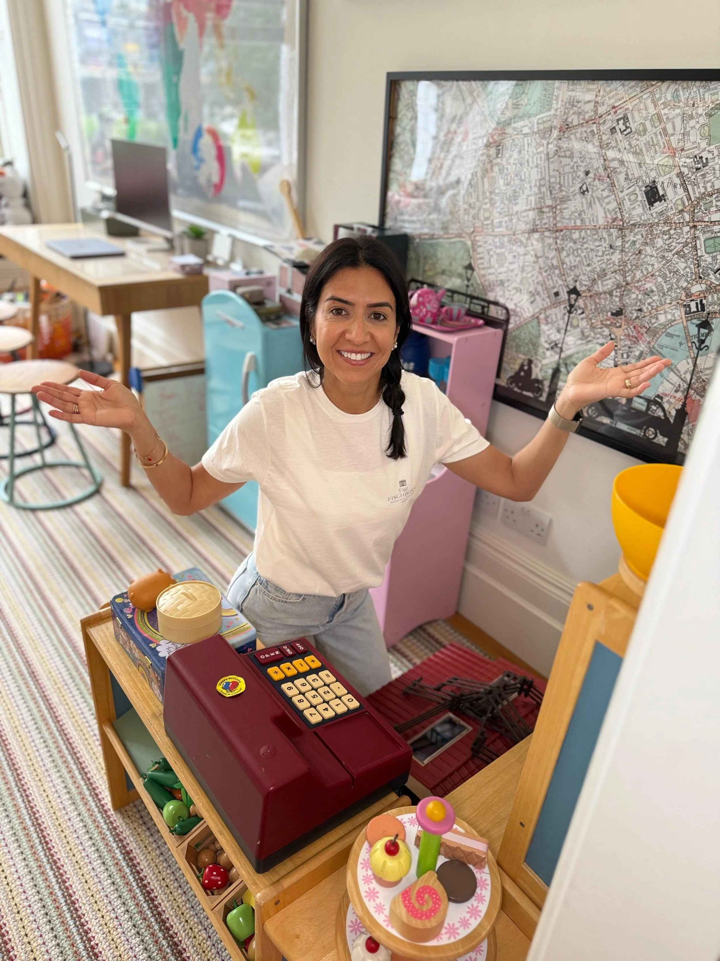 Woman standing behind a toy cash register in a colorful play area, smiling, with a large map on the wall behind her.