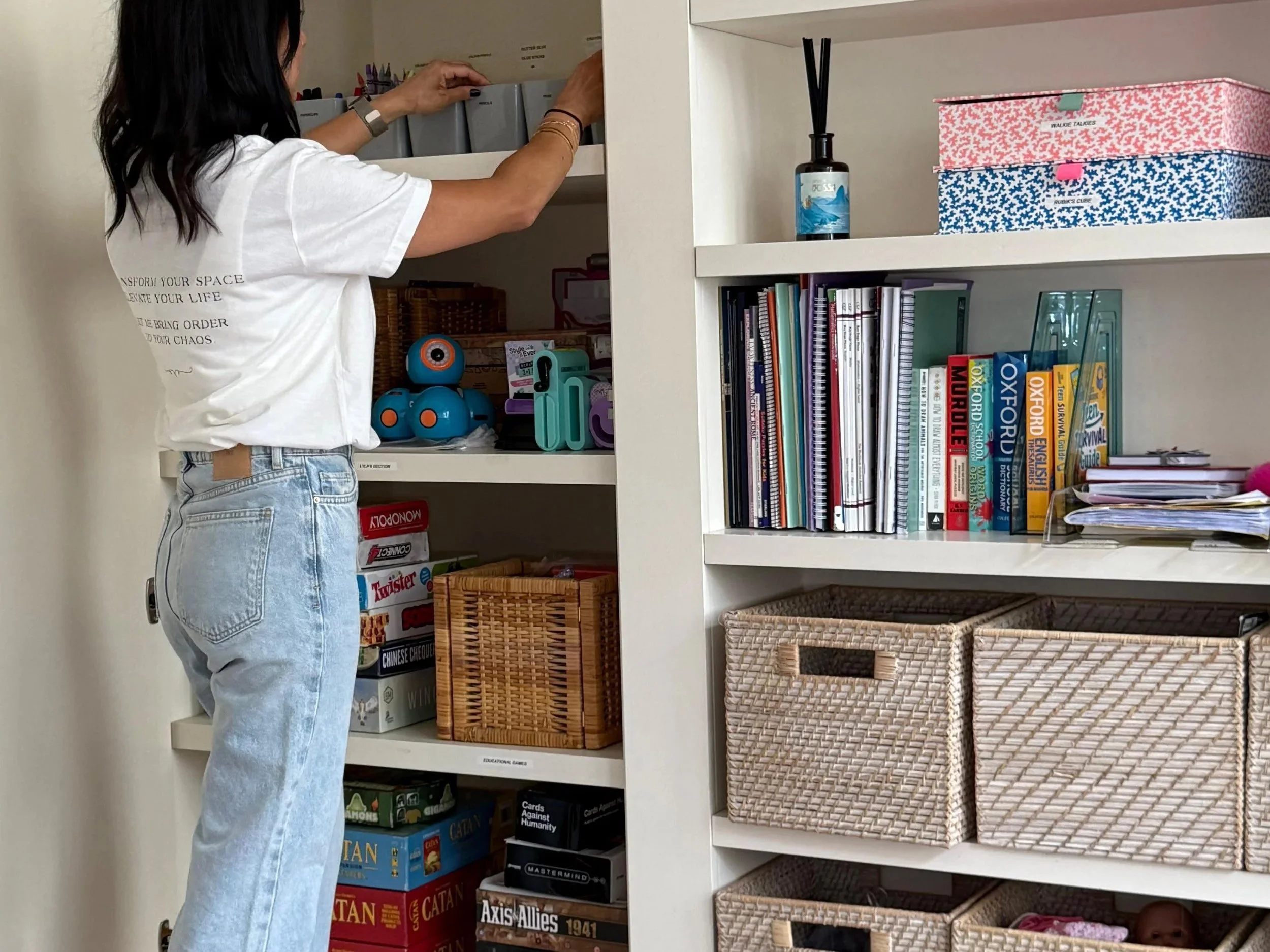A woman organizing shelves in a built-in white bookshelf, which contains books, decorative items, and woven storage baskets in a cozy room with a cream-colored rug and white wall paneling.