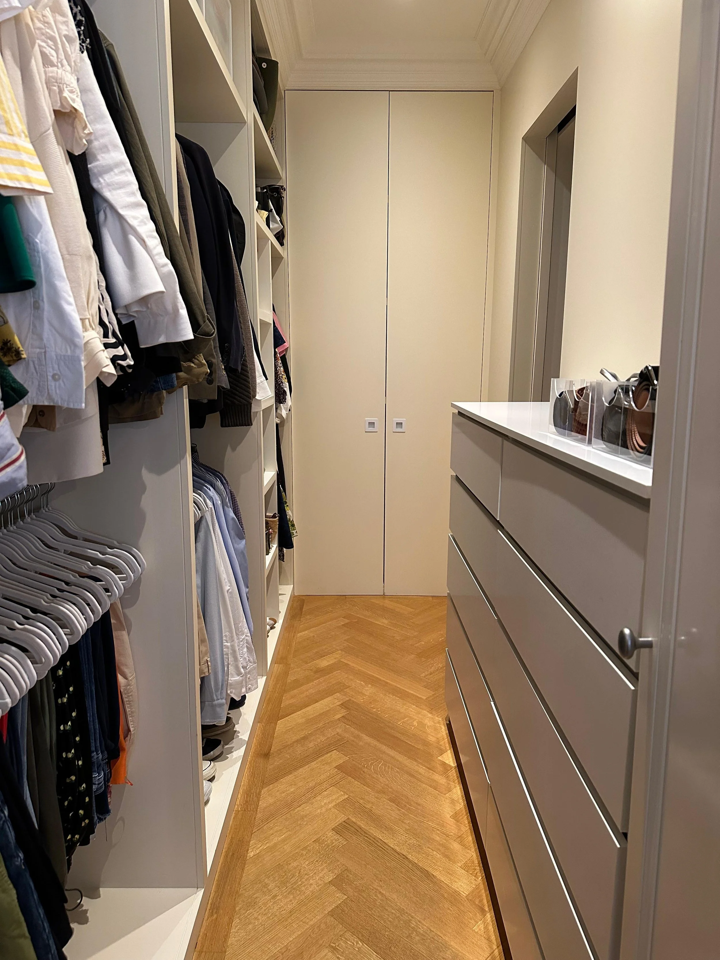 A walk-in closet with white shelving on the left filled with clothes and accessories, a wooden parquet floor, and a white dresser on the right side. There's a closed cabinet at the end of the closet.