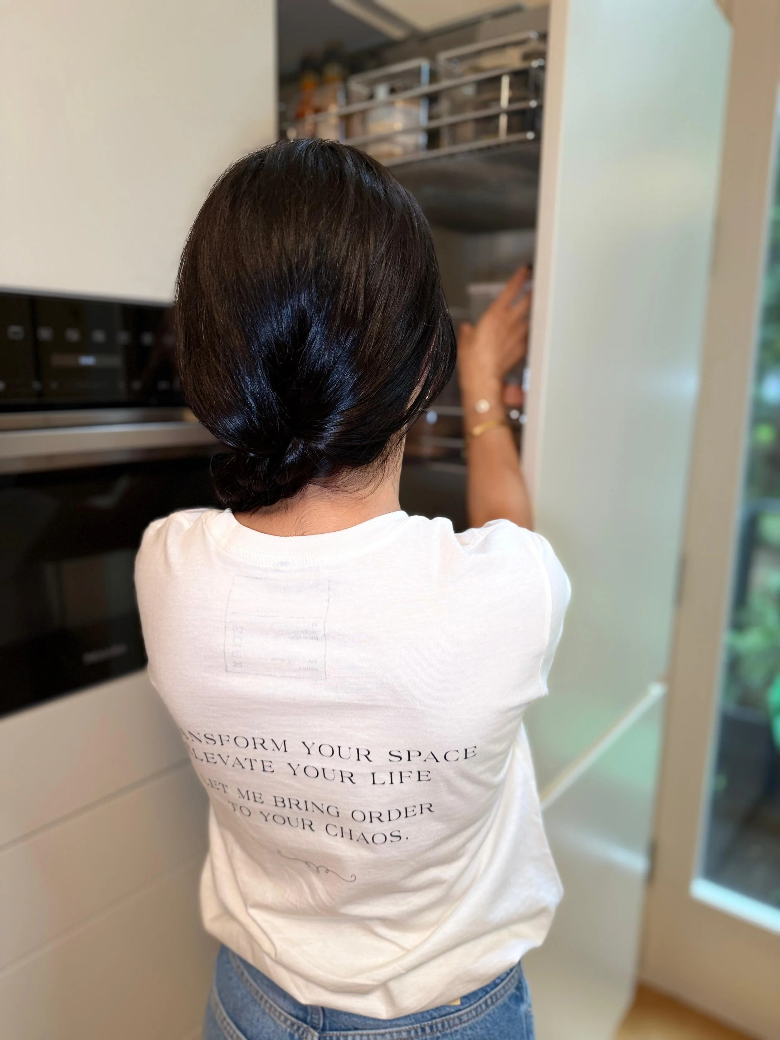 A woman with dark hair styled in a low bun, wearing a white t-shirt, is reaching into a pantry or closet with metal shelves filled with various items.