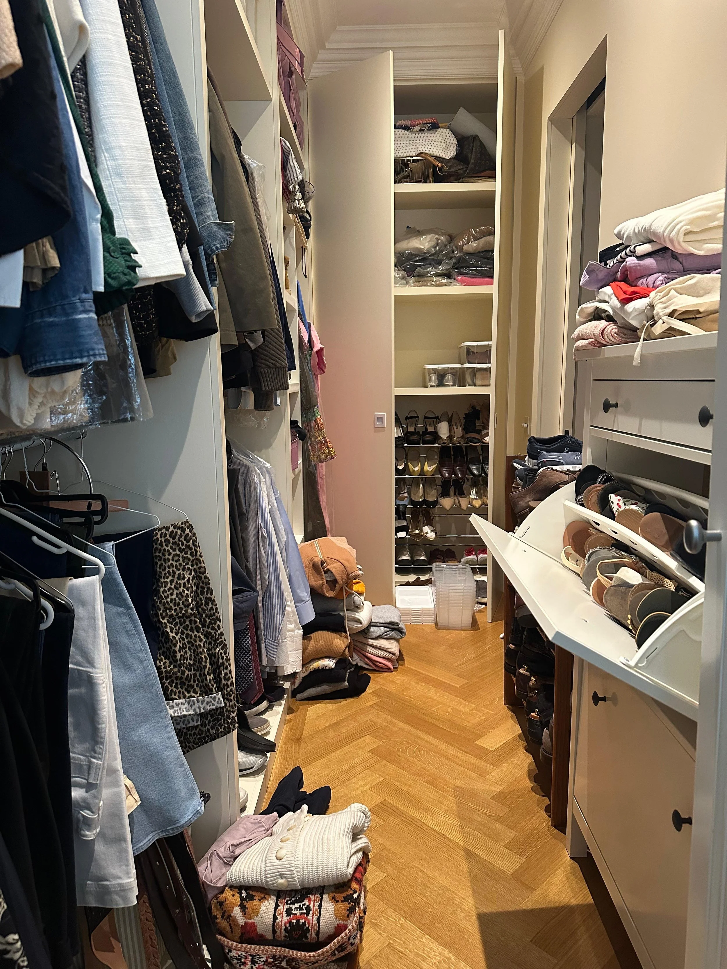 A walk-in closet with clothes hanging on the left, shoes on multiple shelves and racks at the back, and various folded clothes on shelves and surfaces on the right.