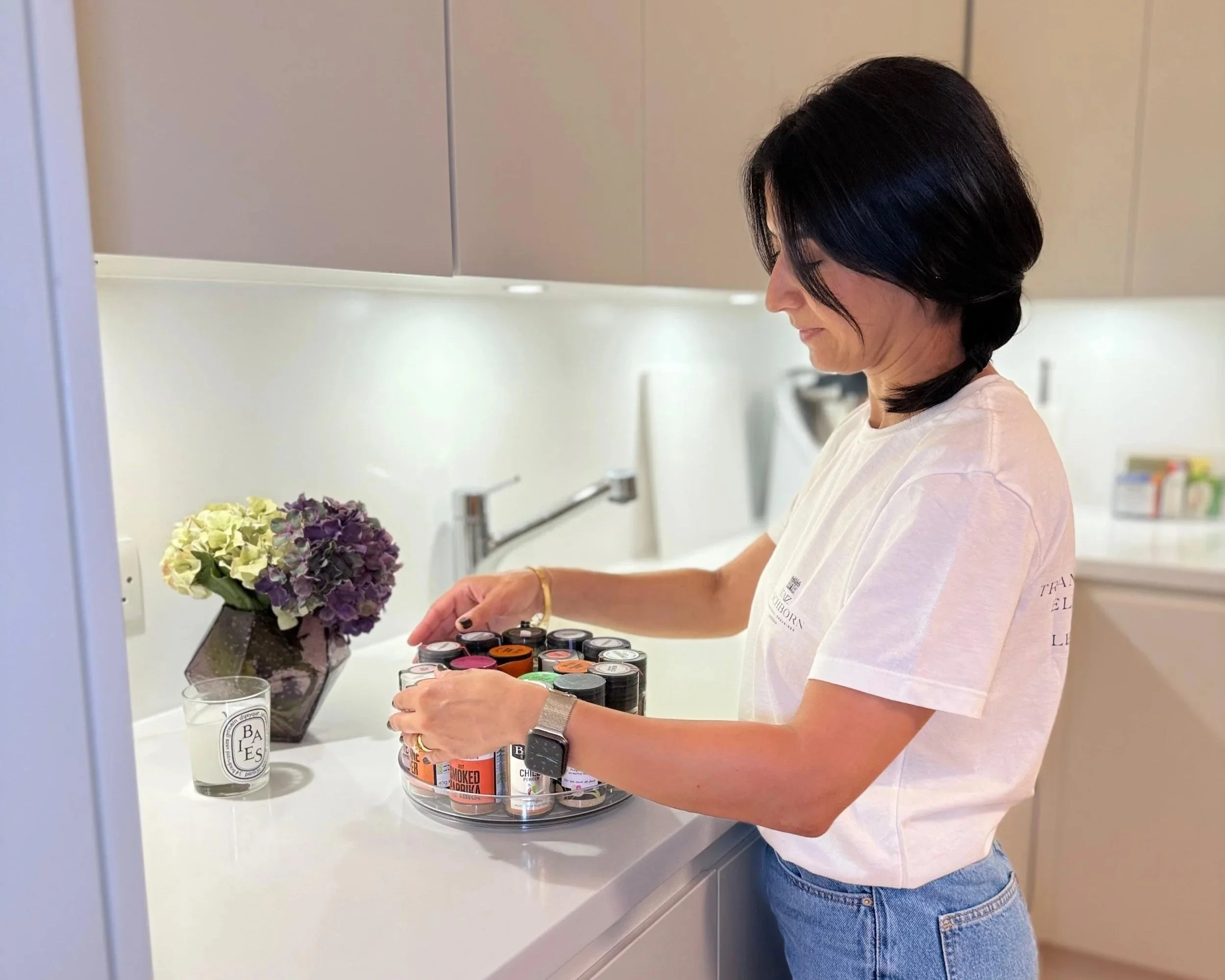A woman with shoulder-length black hair is organizing a tray of various small cans and jars in a kitchen.