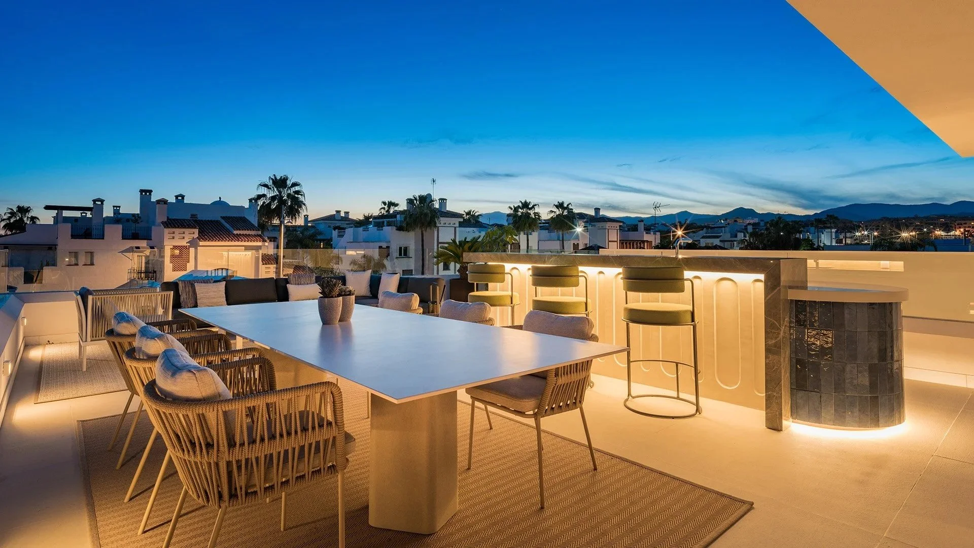 Rooftop patio with dining table, chairs, and barstools against a backdrop of a sunset over a city with palm trees and mountains in the distance.