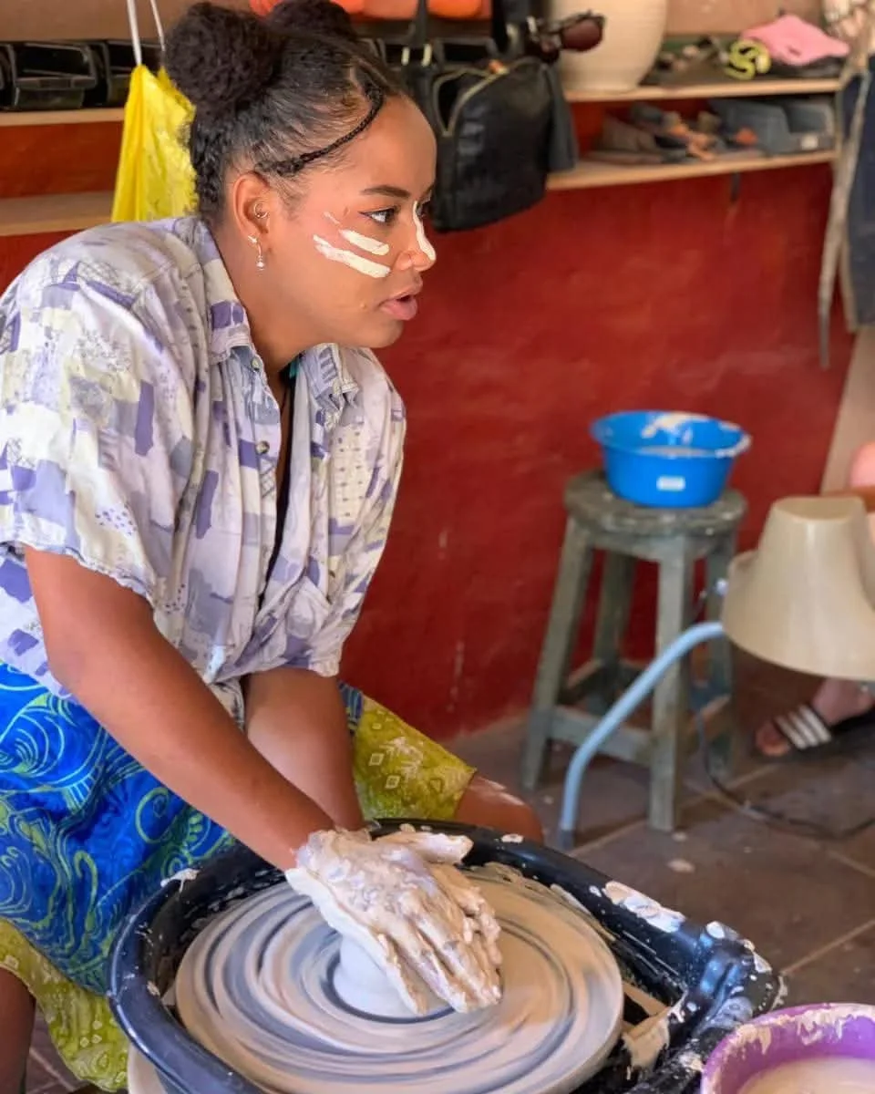 A young woman with face paint on her cheeks is shaping clay on a pottery wheel. She has dark hair tied up and is wearing a patterned shirt and colourful shorts. The background shows shelves with pottery supplies and tools.