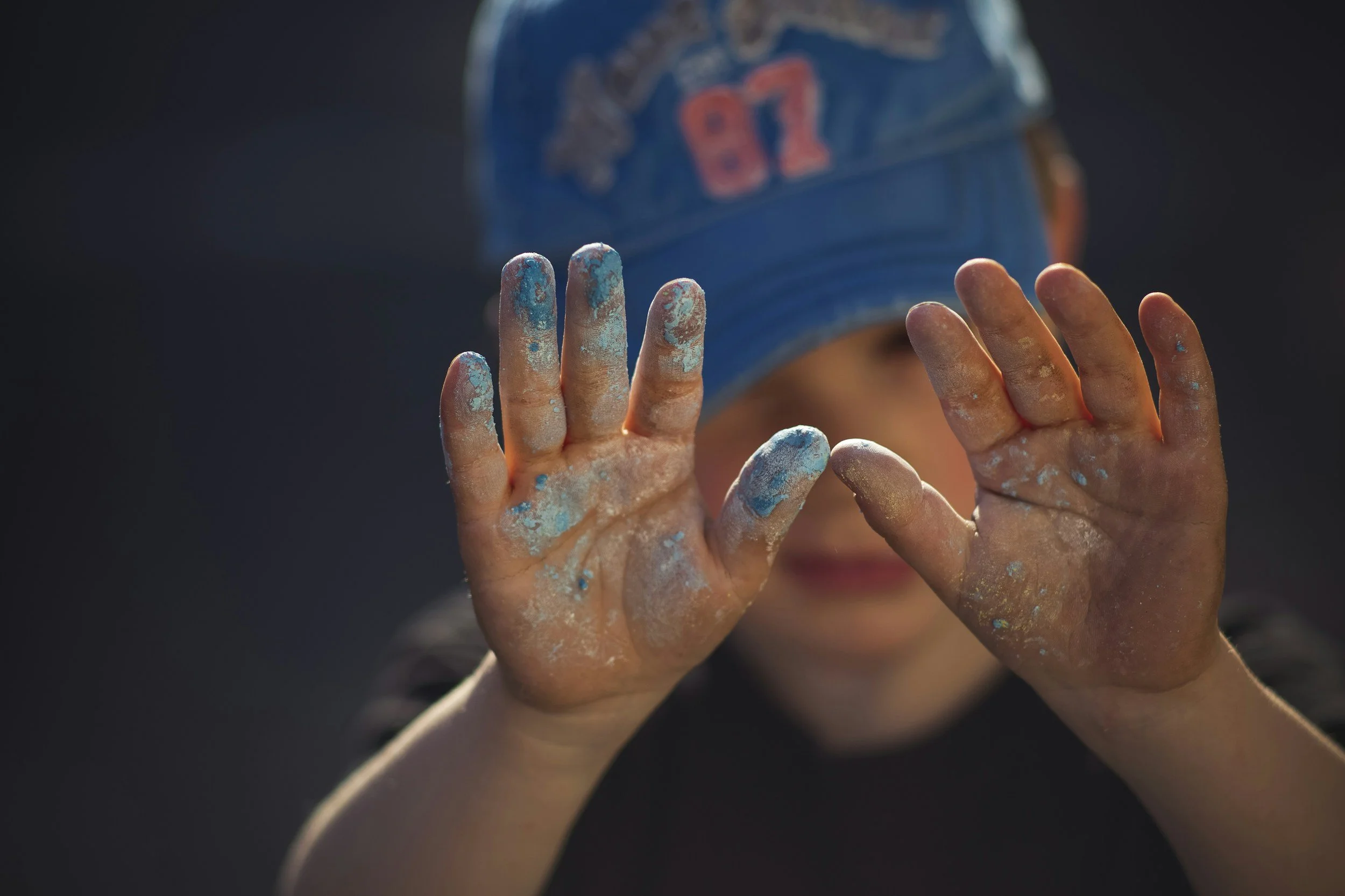 A young child with dirty hands covered in blue and white paint, holding up both hands with fingers spread apart, wearing a blue baseball cap and a dark shirt.