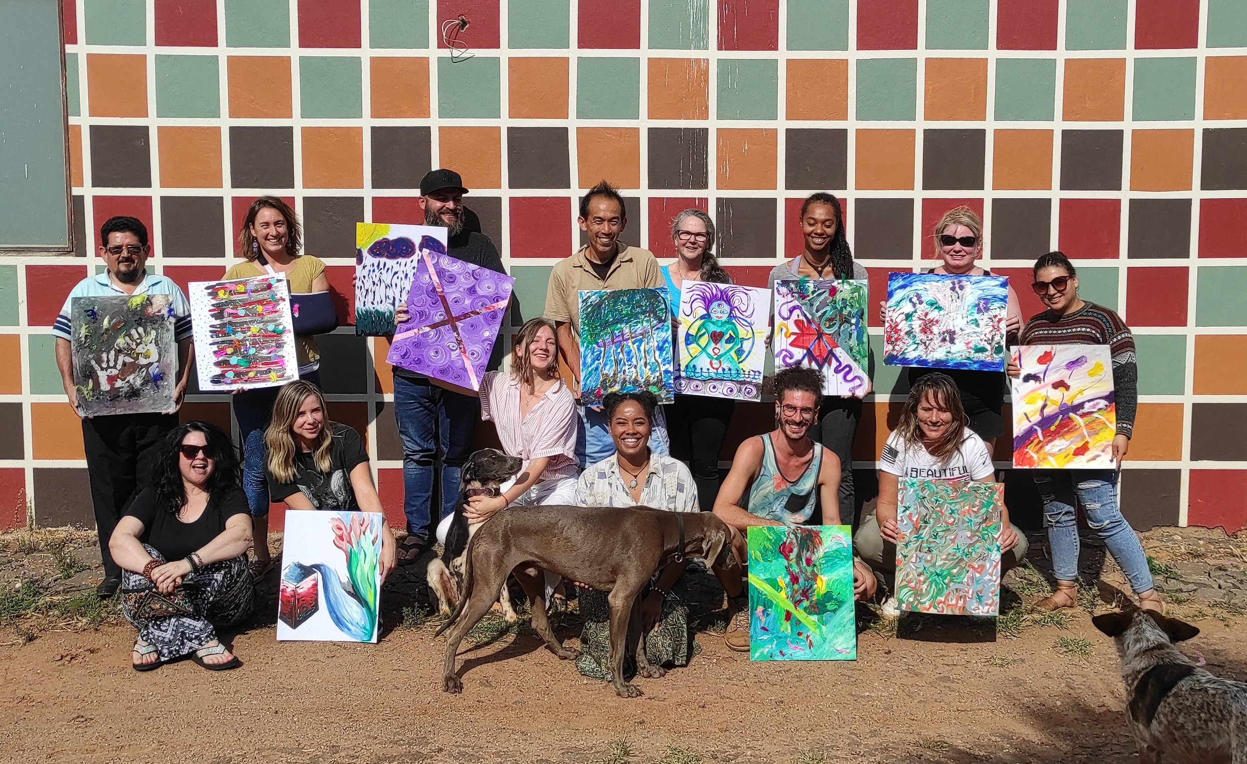 A group of fifteen people holding colourful abstract paintings in front of a multicoloured tiled wall, with two dogs in the foreground, one sitting and one standing, outdoors.
