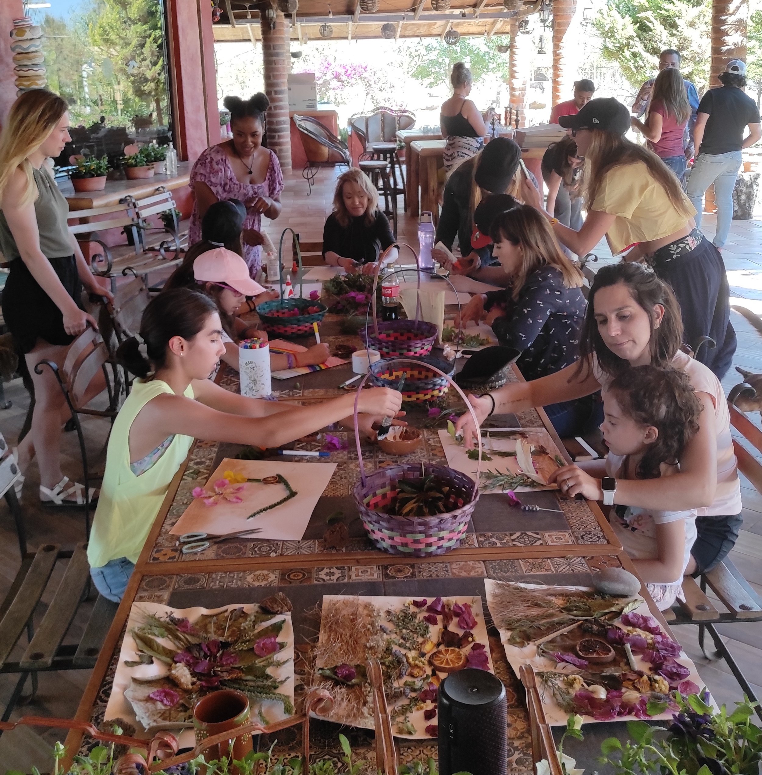 A group of children and adults participating in a flower arranging workshop at a long table covered with flower supplies, in a brightly lit outdoor space with large windows showing greenery outside.