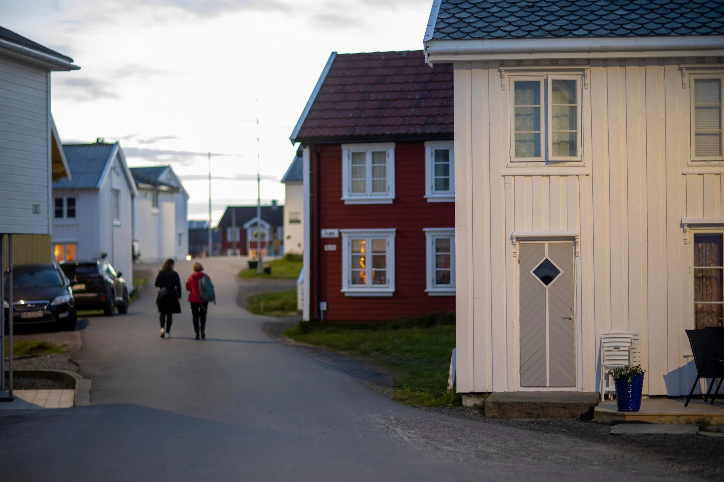 A street scene in a neighborhood with colorful houses, two women walking on the sidewalk, parked cars, and a cloudy sky.