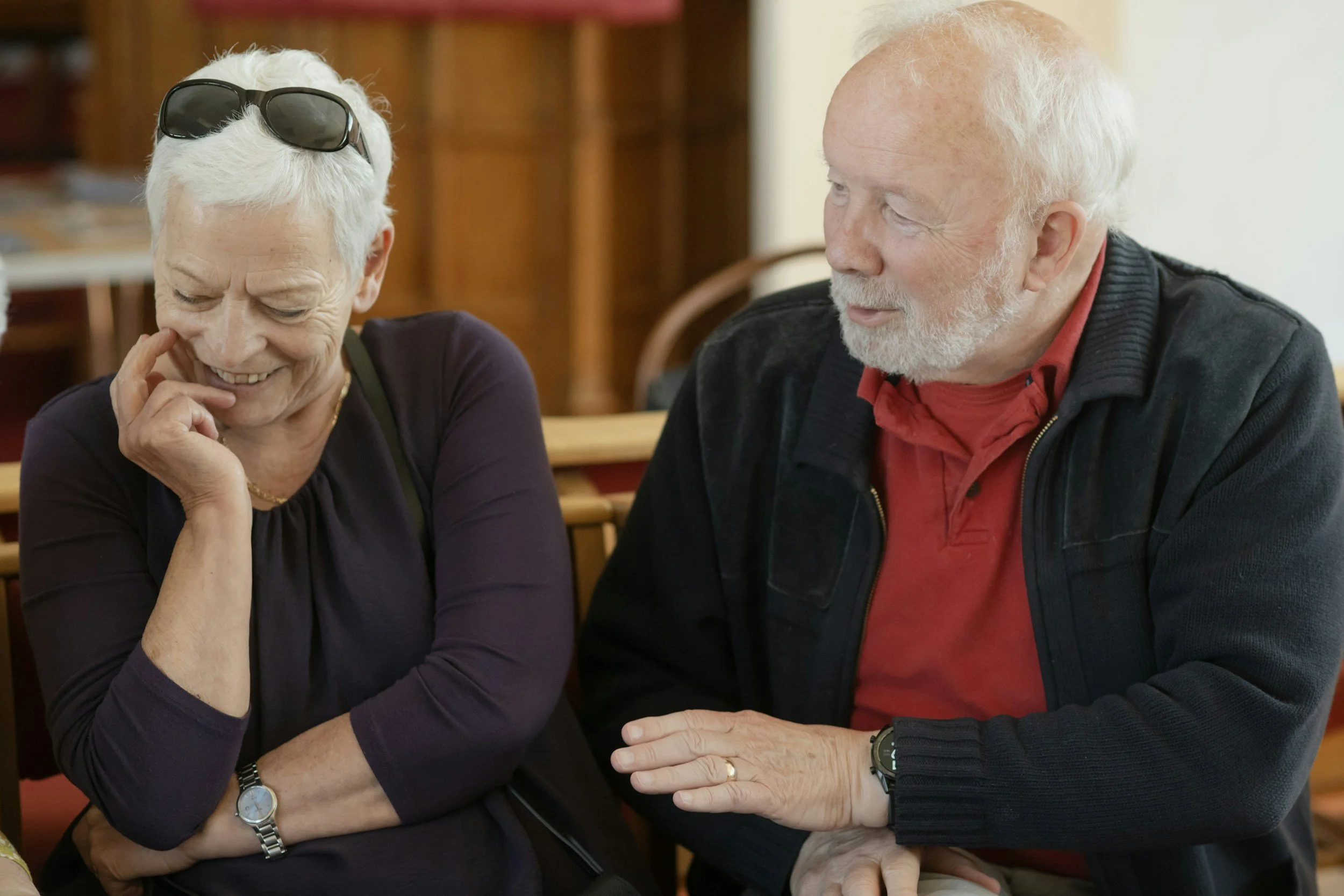 An elderly man and woman sitting together, sharing a moment of laughter. The woman has white hair with sunglasses on her head, and the man has white hair and a beard. They are sitting at a table in a cozy, warmly lit setting.