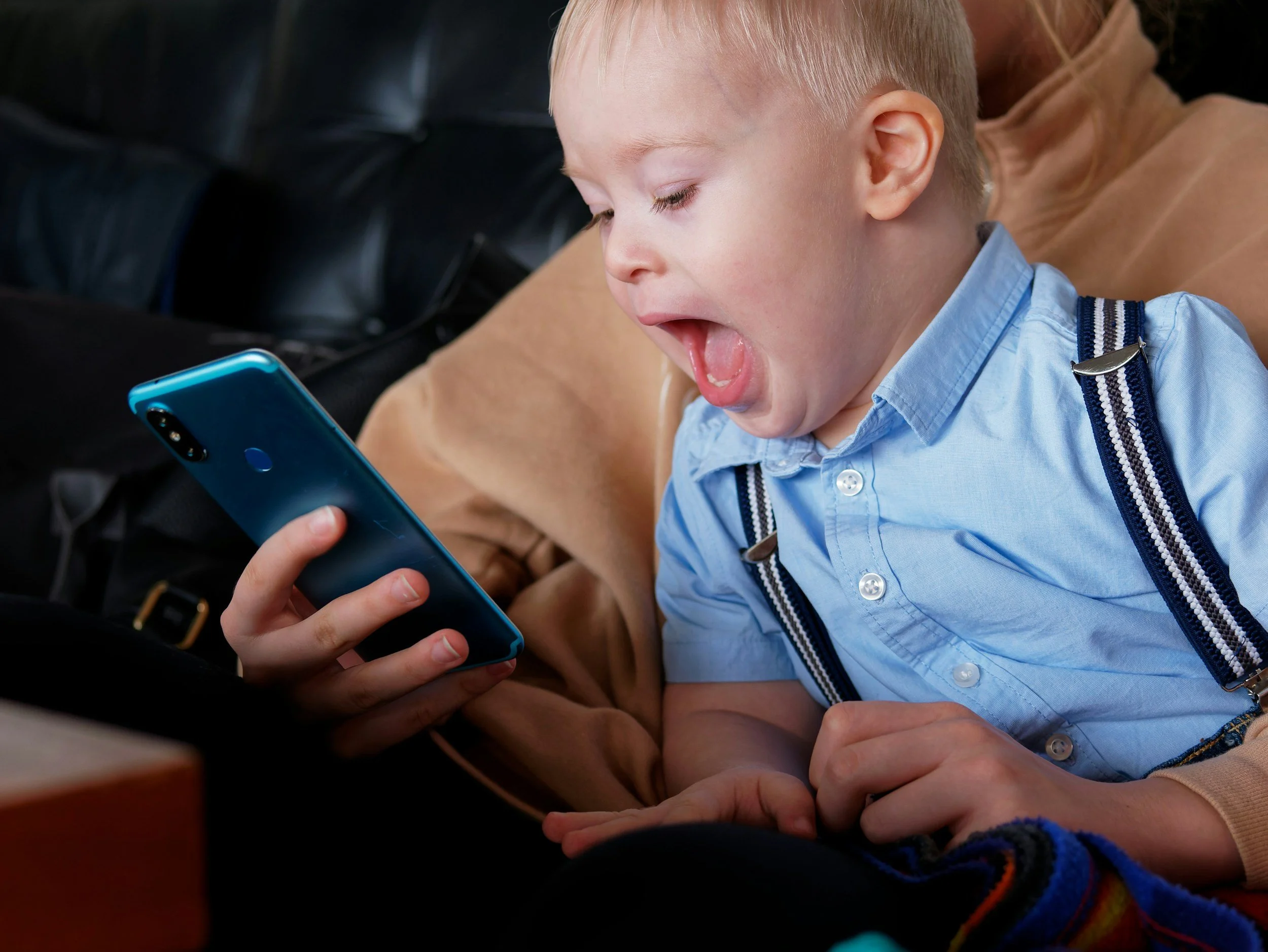 A young boy with blond hair wearing a blue shirt with suspenders, looking surprised and engaged as he looks at a smartphone held by an adult with a watch on their wrist.